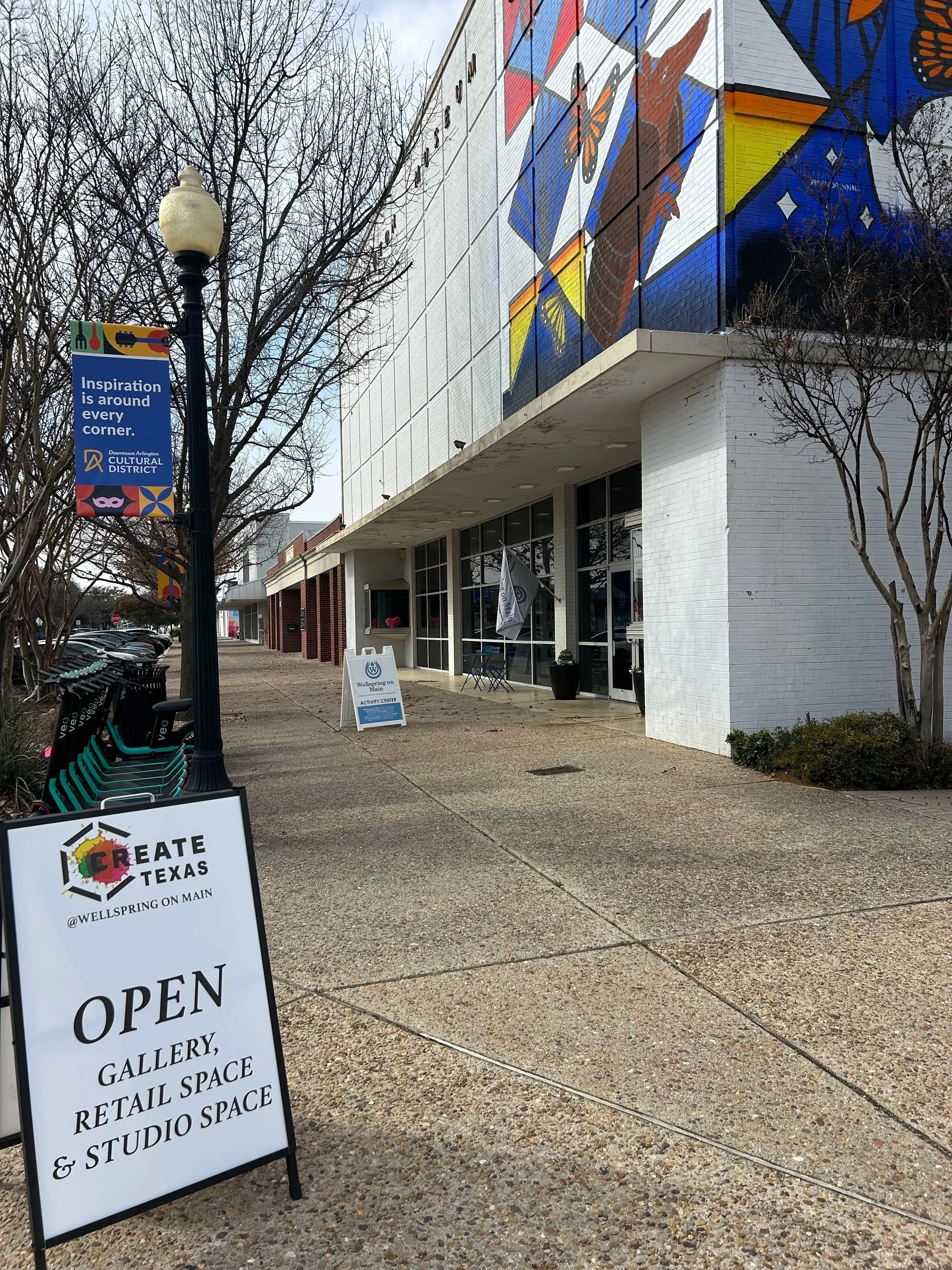 Empty sidewalk outside Create Texas art gallery and retail space with a signboard indicating it is open, and a mural of a woman with a computer on the building wall.