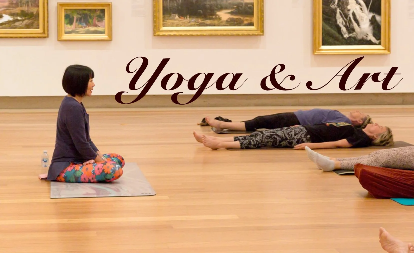 An indoor yoga class in a gallery with paintings on the wall. A woman sits cross-legged on a yoga mat facing two people lying on their backs on the floor in Savasana pose. The text 'Yoga & Art' is overlaid on the image.