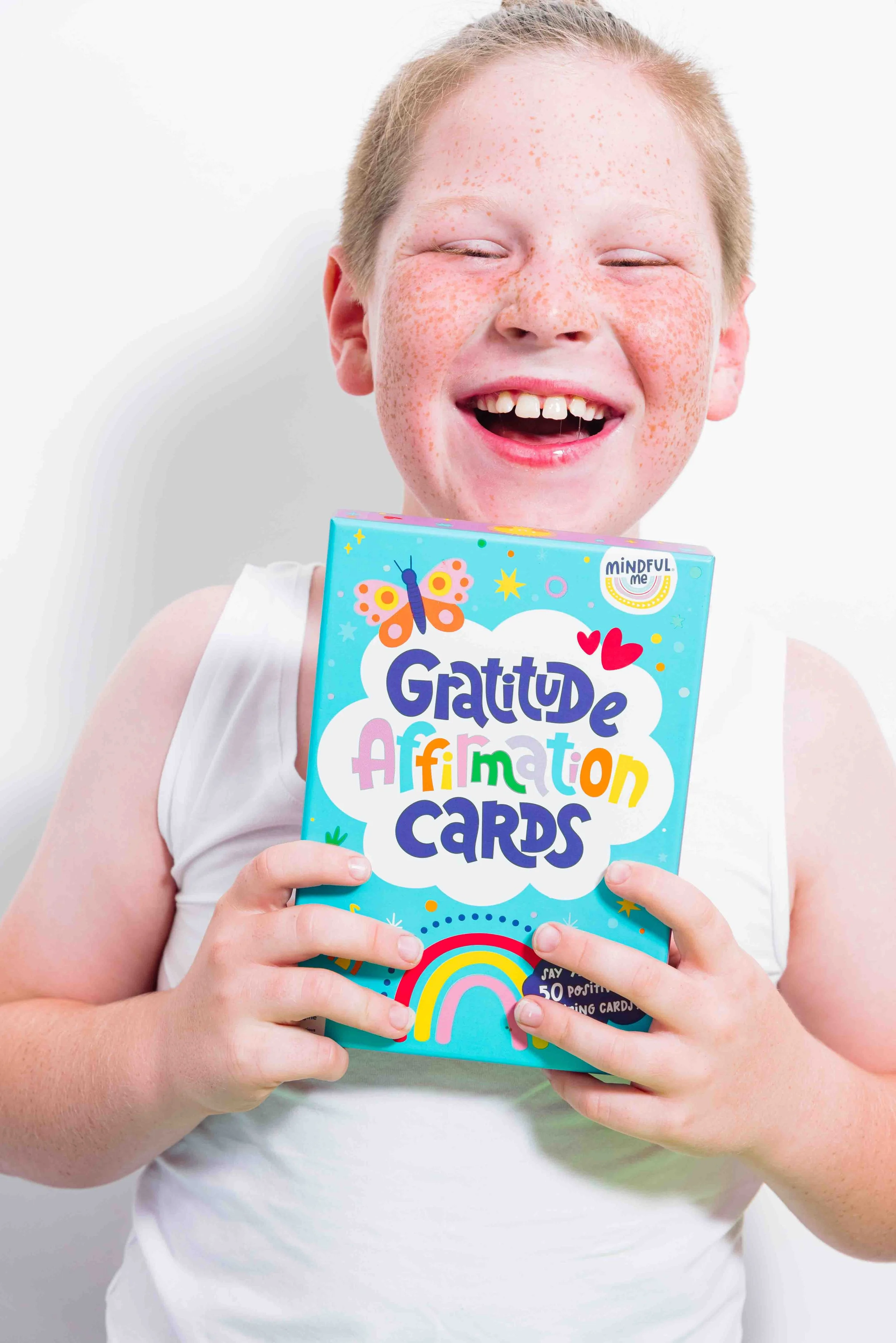 A smiling child with freckles holding a colorful box of gratitude affirmation cards.