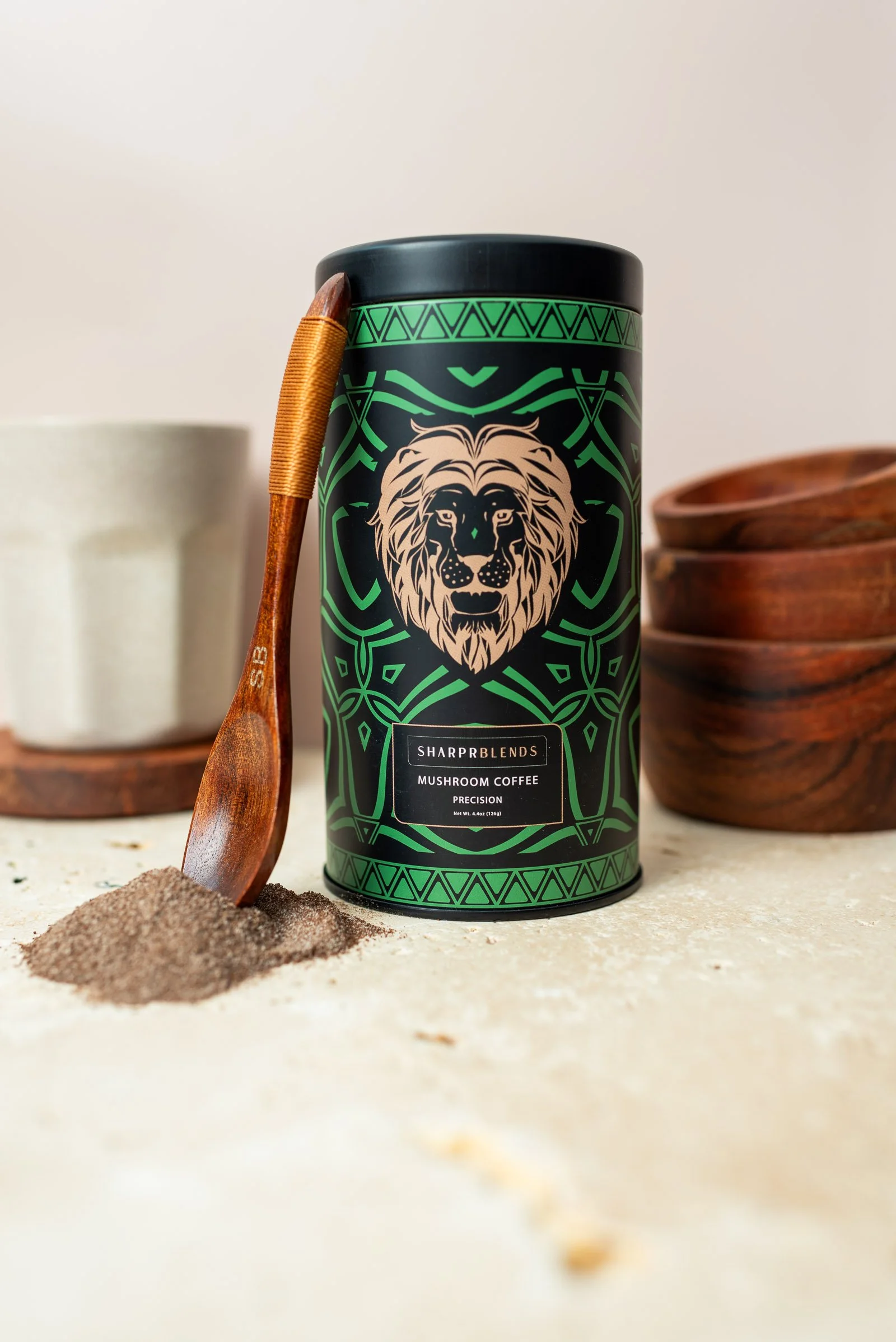 A black container labeled 'Sharp Blends Mushroom Coffee' with a lion illustration, placed on a light surface next to a wooden spoon with some ground mushroom coffee spilled on it and wooden bowls in the background.