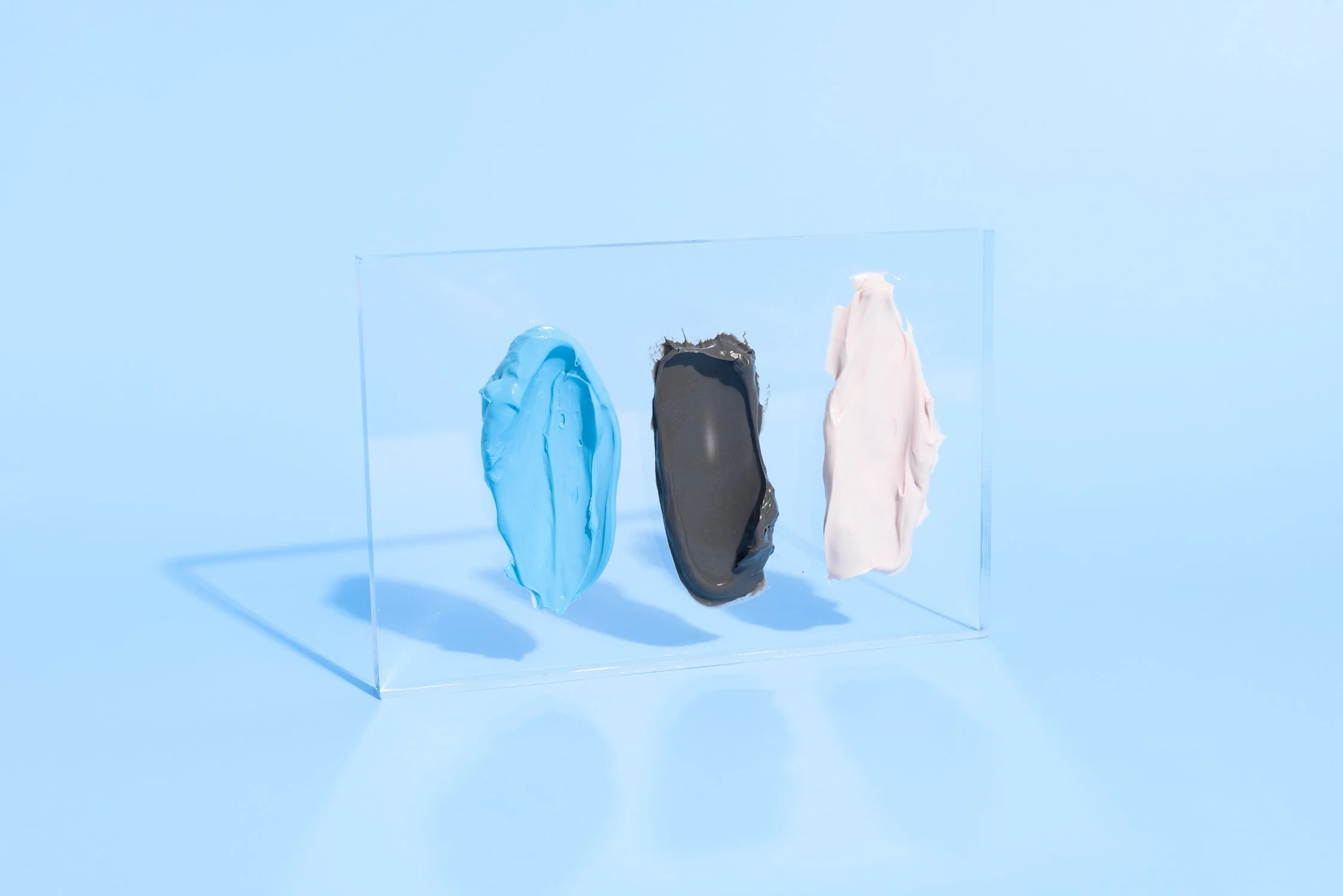 Three blobs of paint in blue, black, and pink are displayed on a clear acrylic panel against a light blue background.