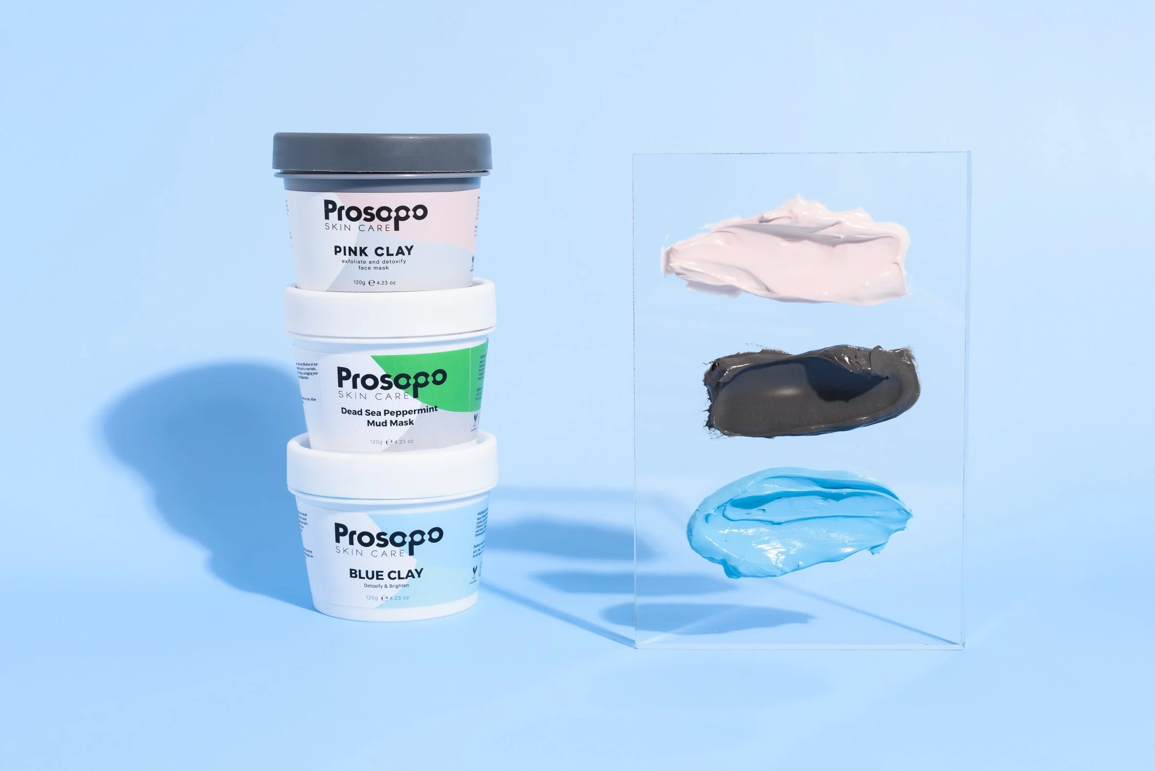 Three containers of Prospa skincare masks stacked vertically next to a transparent glass panel with swatches of pink, black, and blue clay masks on it. The background is light blue.