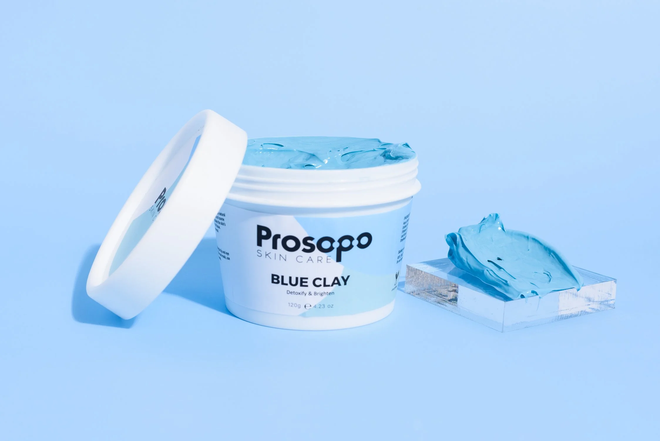 A white container of Prospo Blue Clay skincare product with the lid off, showing blue clay inside. A clear acrylic display with blue clay on it is beside the container, set against a blue background.