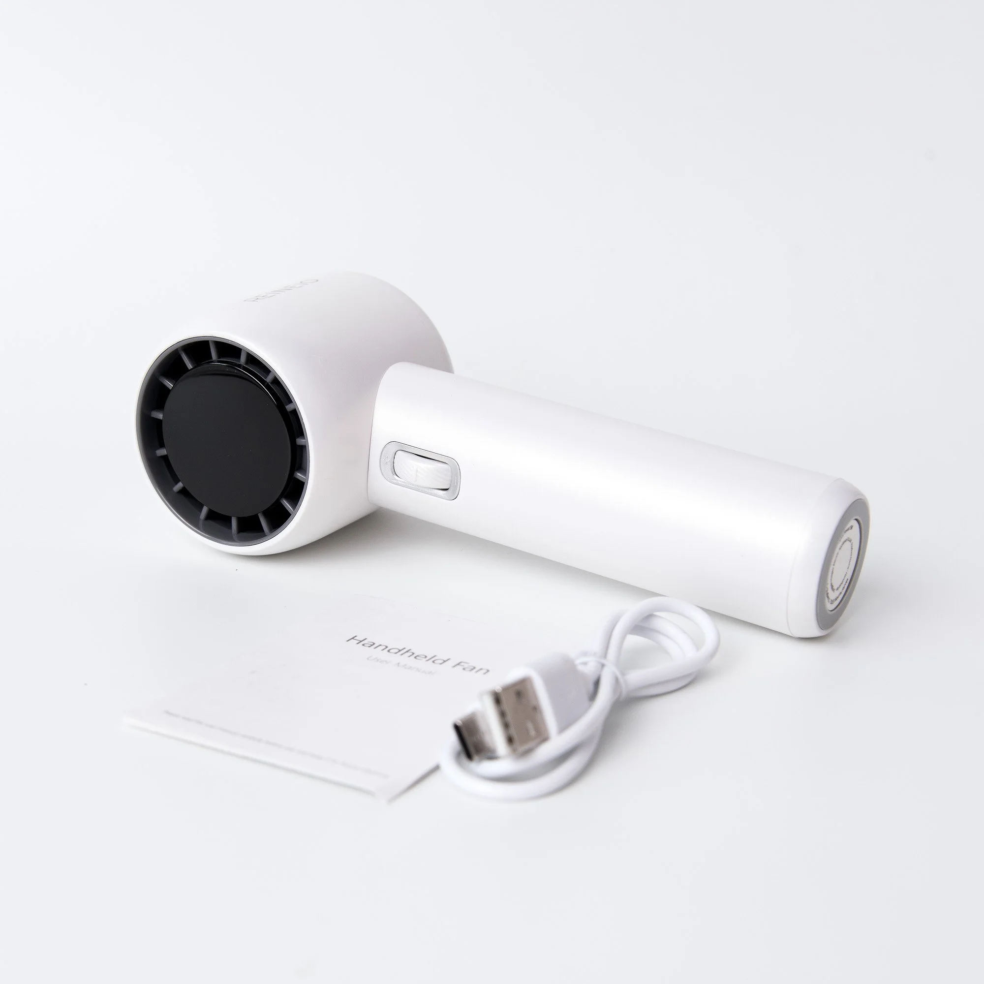 White handheld fan with a circular black vent, a power button on the handle, a USB charging cable, and user manual labeled 'Handheld Fan' on a white background.