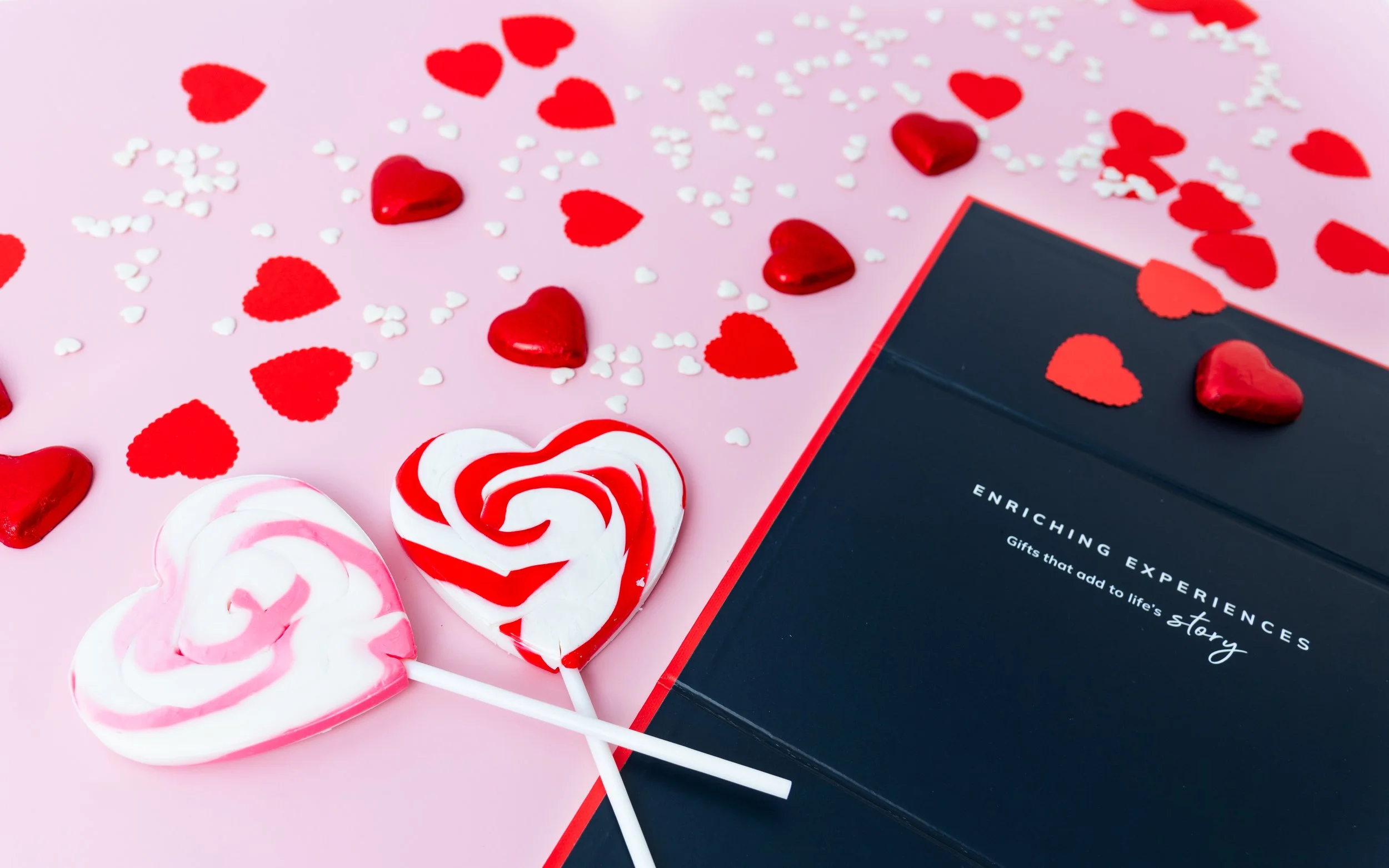 Two heart-shaped lollipops, one pink and white and the other red and white, placed on a pink surface with scattered red and white heart and circle-shaped confetti. A dark-colored box with the text 'Enriching Experiences Gifts that add to life's story