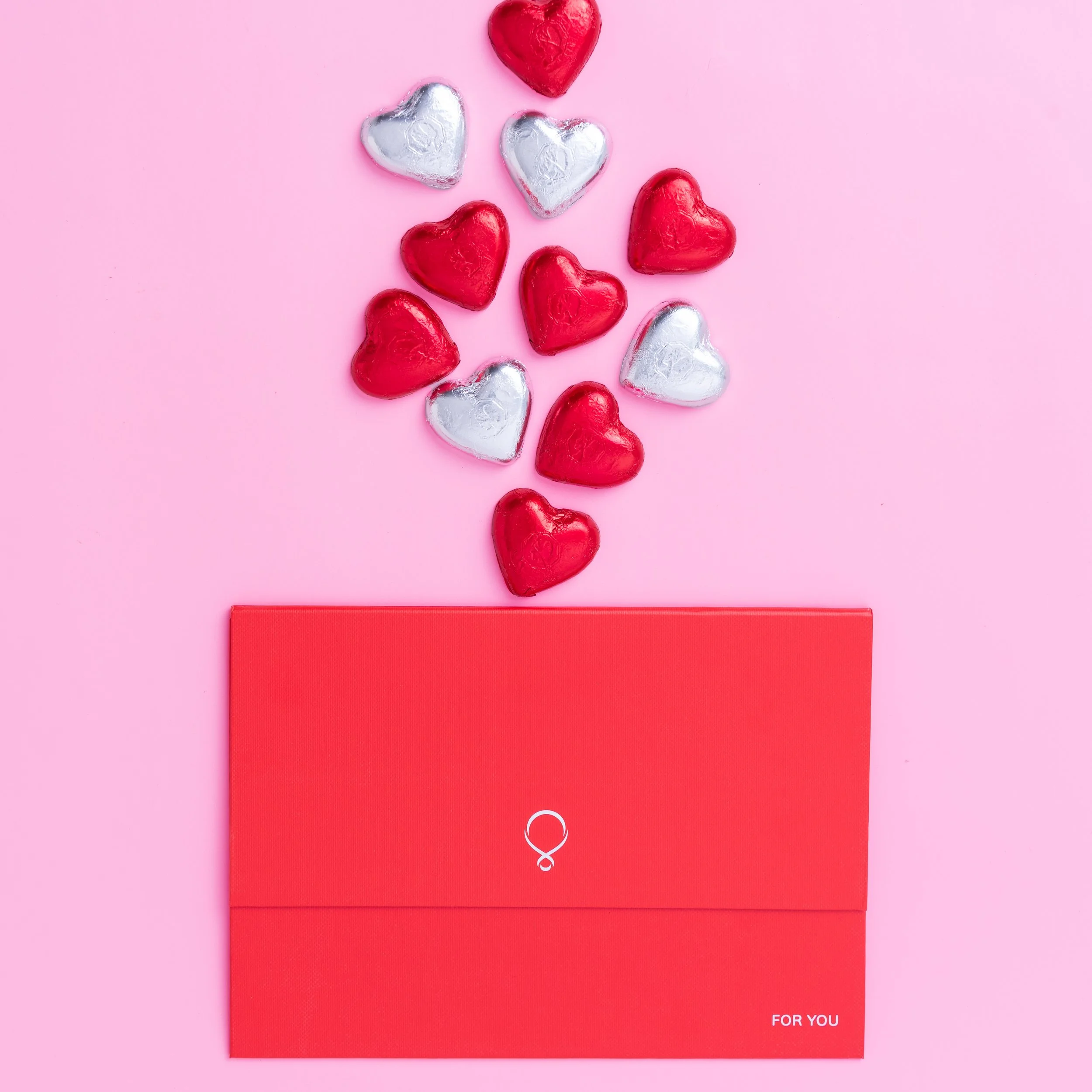 Red and silver foil-wrapped heart chocolates spilling out from a red envelope marked 'For You' on a pink background.