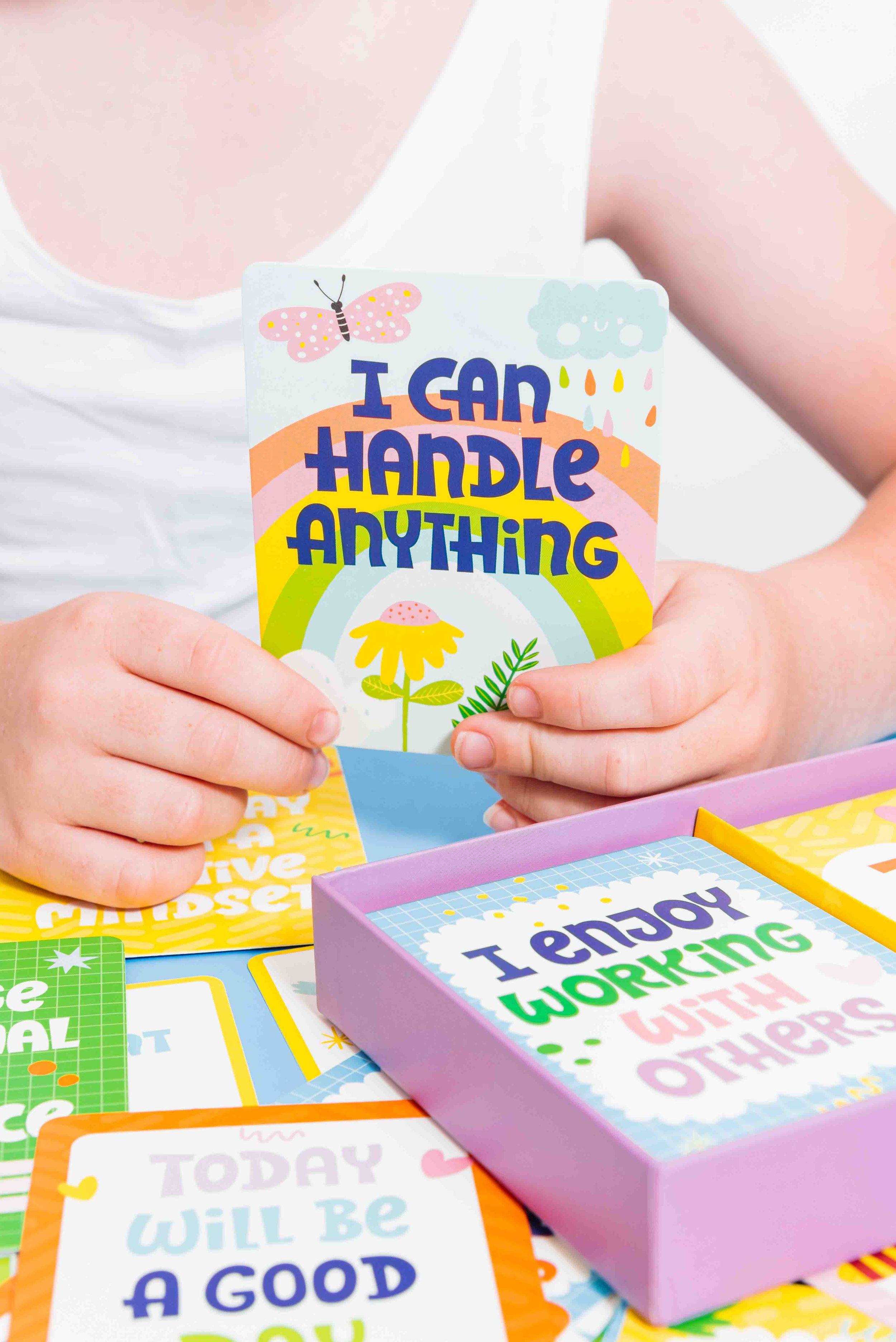Person holding a colorful card that says 'I can handle anything,' with rainbow, cloud, butterfly, and floral illustrations, surrounded by other motivational cards on a table.