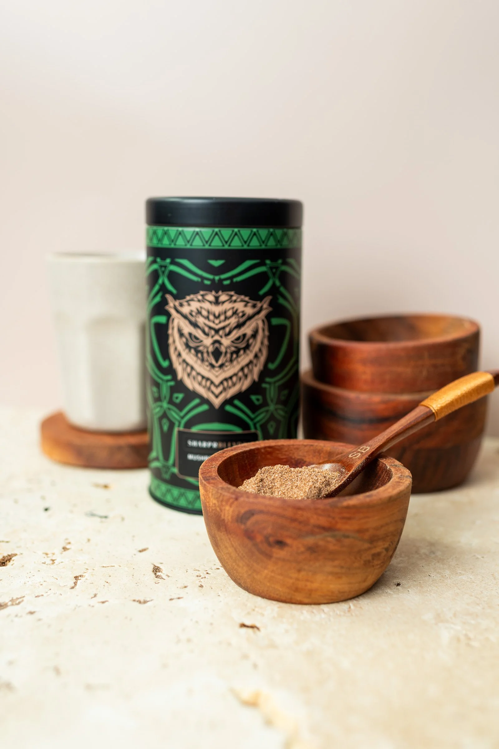 A black and green container with an owl illustration, a small white cup, and three wooden bowls with a wooden spoon in one bowl, on a marble surface.
