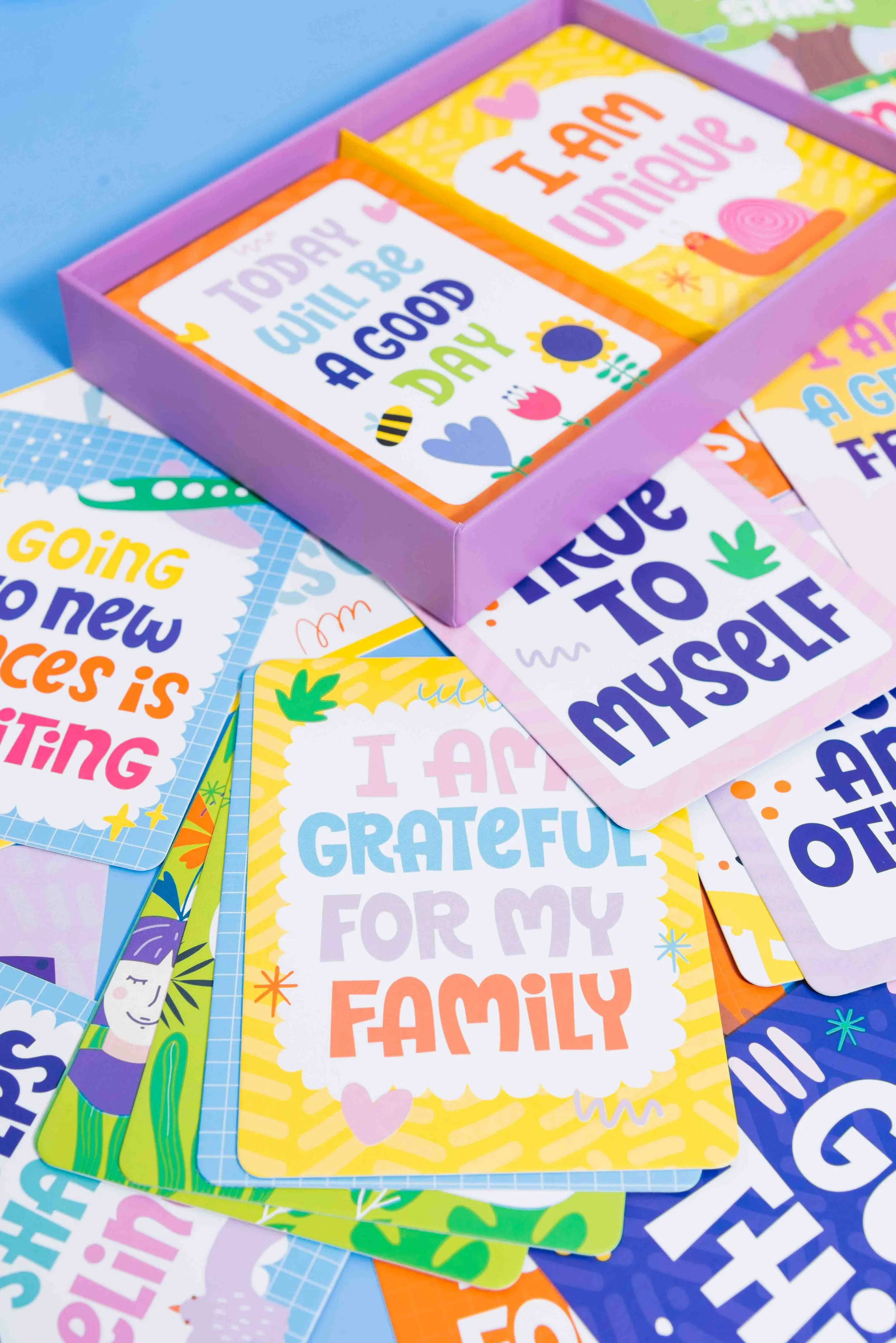 Colorful motivational cards with positive messages, some stacked and some arranged in a purple box, featuring phrases like 'Today will be a good day,' 'I am grateful for my family,' and 'I am unique.'