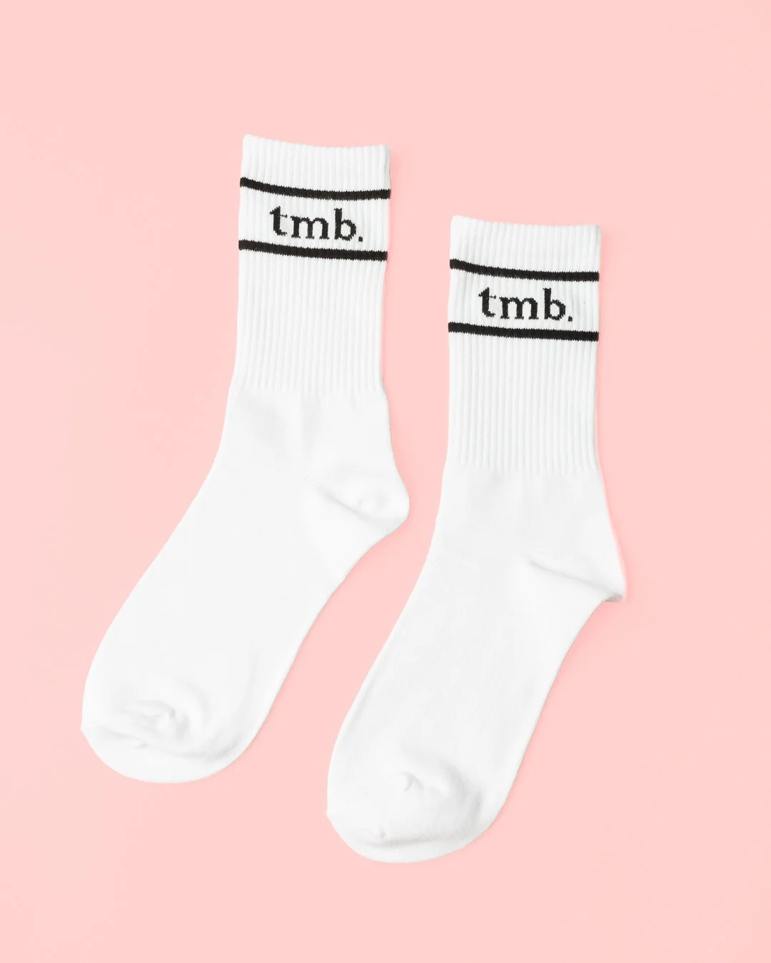 A pair of white crew socks with black stripes and the text "tmb." on the cuff, set against a pink background.