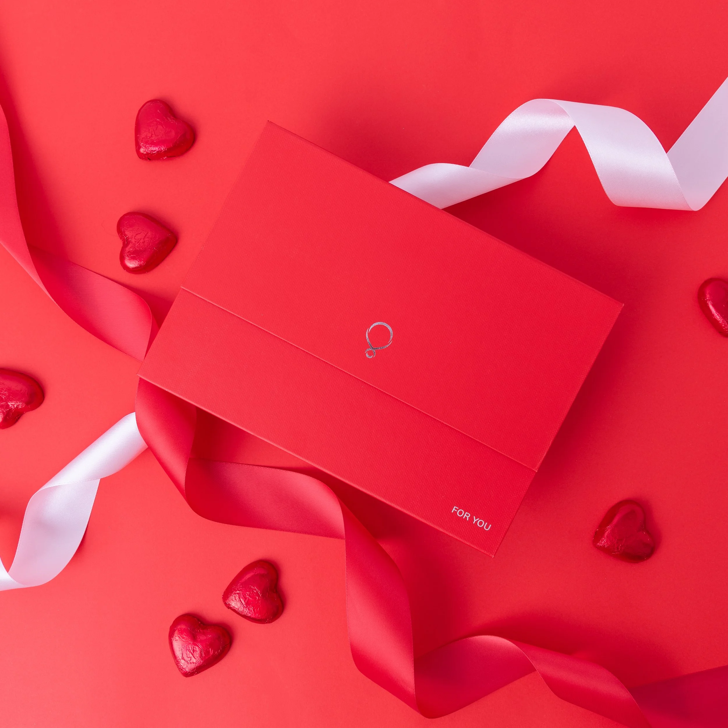 Red gift box with white ribbon on a red background, surrounded by small red foil-wrapped hearts, with a small silver ring illustration on the box.