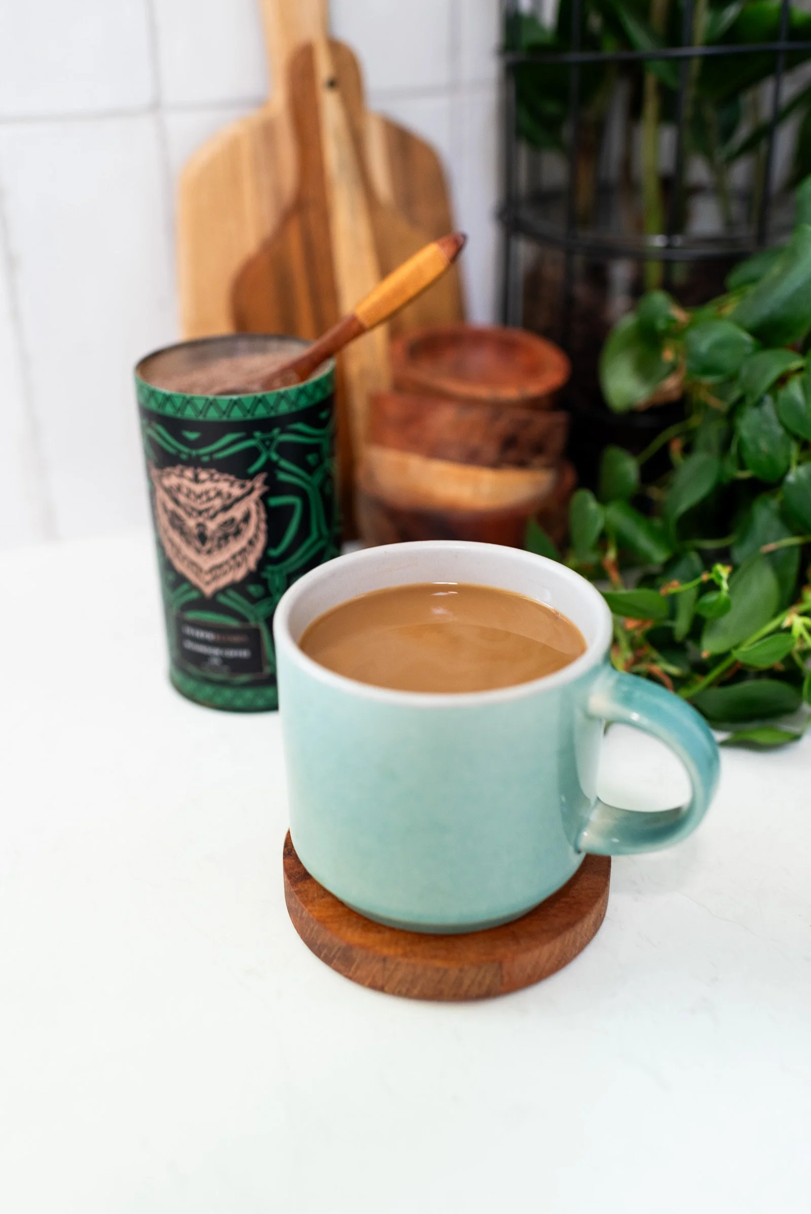 A light blue ceramic mug filled with coffee on a wooden coaster, placed on a white surface. Behind it are a green and black travel mug with a straw and a decorative owl design, wooden bowls, and green plants in a black wire holder, all in a kitchen s