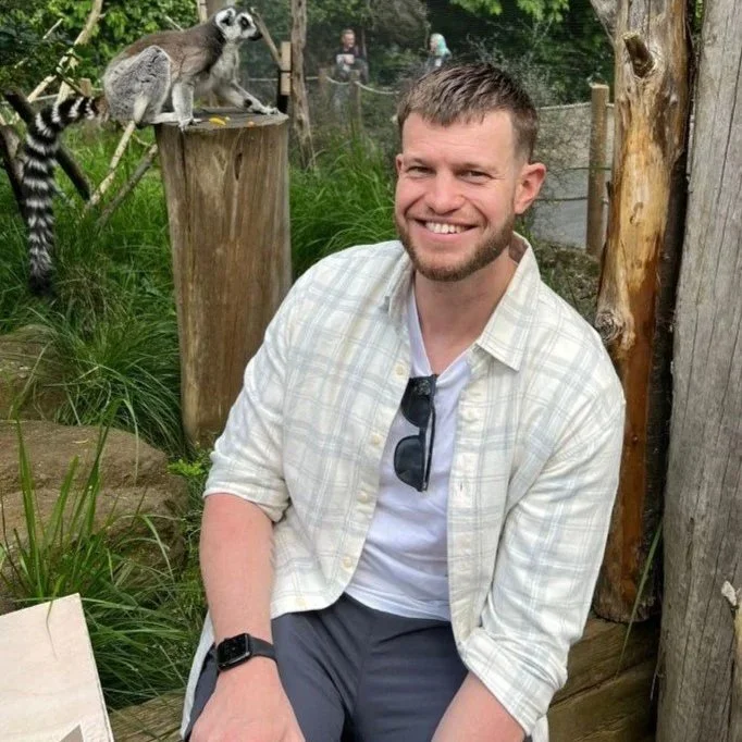 Volunteer mentor Oliver sat smiling infront of a lemur enclosure.