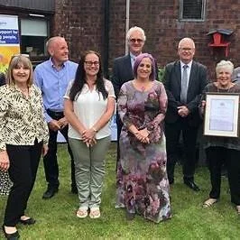  Seven adults, standing in front of a brick building on the grass, dressed in suits or smart top and trousers, or a dress. One is holding the certificate to show the Queens Award 
