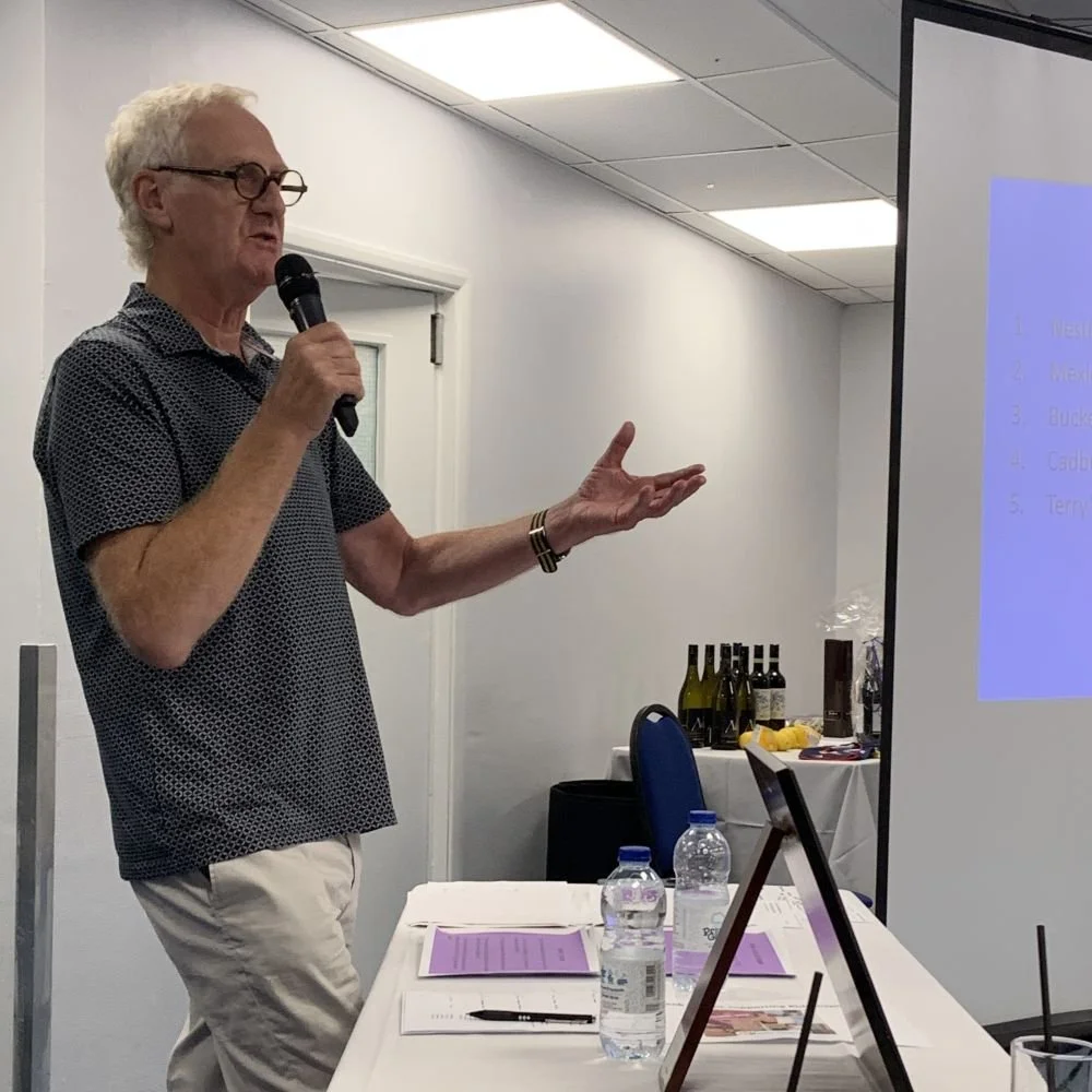  Dave, wearing pale trousers and a blue short sleeve polo shirt, holding a mic in his right hand and standing behind a table, compering the 2023 business quiz 
