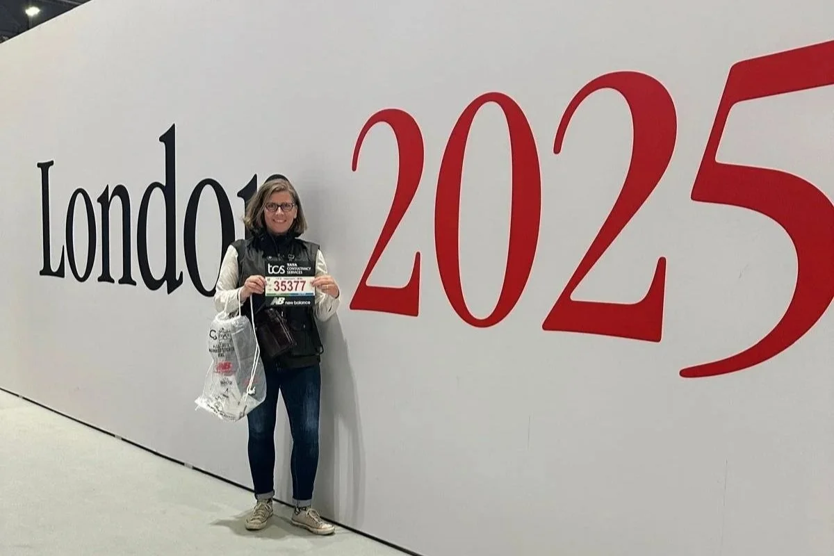 A woman standing with her London Marathon runner number beside a big London Marathon wall.