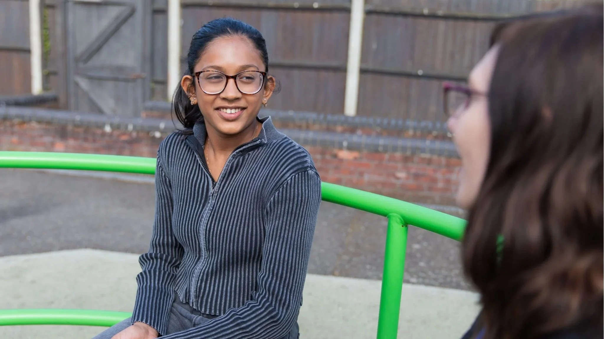 A teenager sat on a roundabout smiling at a Kids Inspire therapist who is sat slightly half in view.