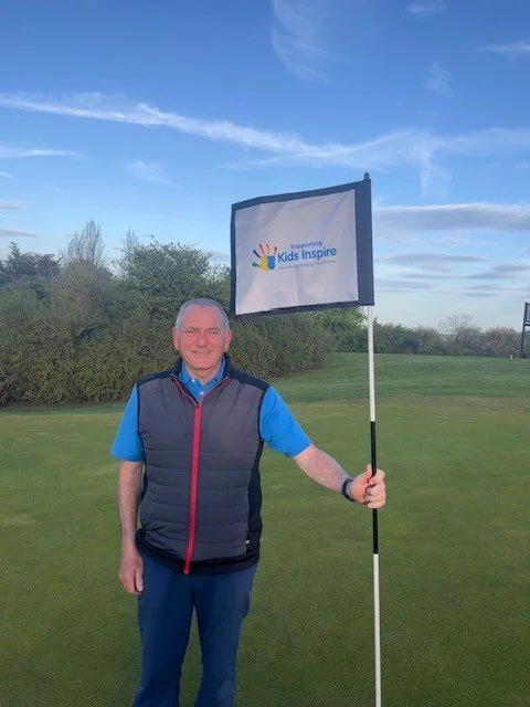 Man stands proudly on a golf course holding a "supporting Kids Inspire" fundraising flag.