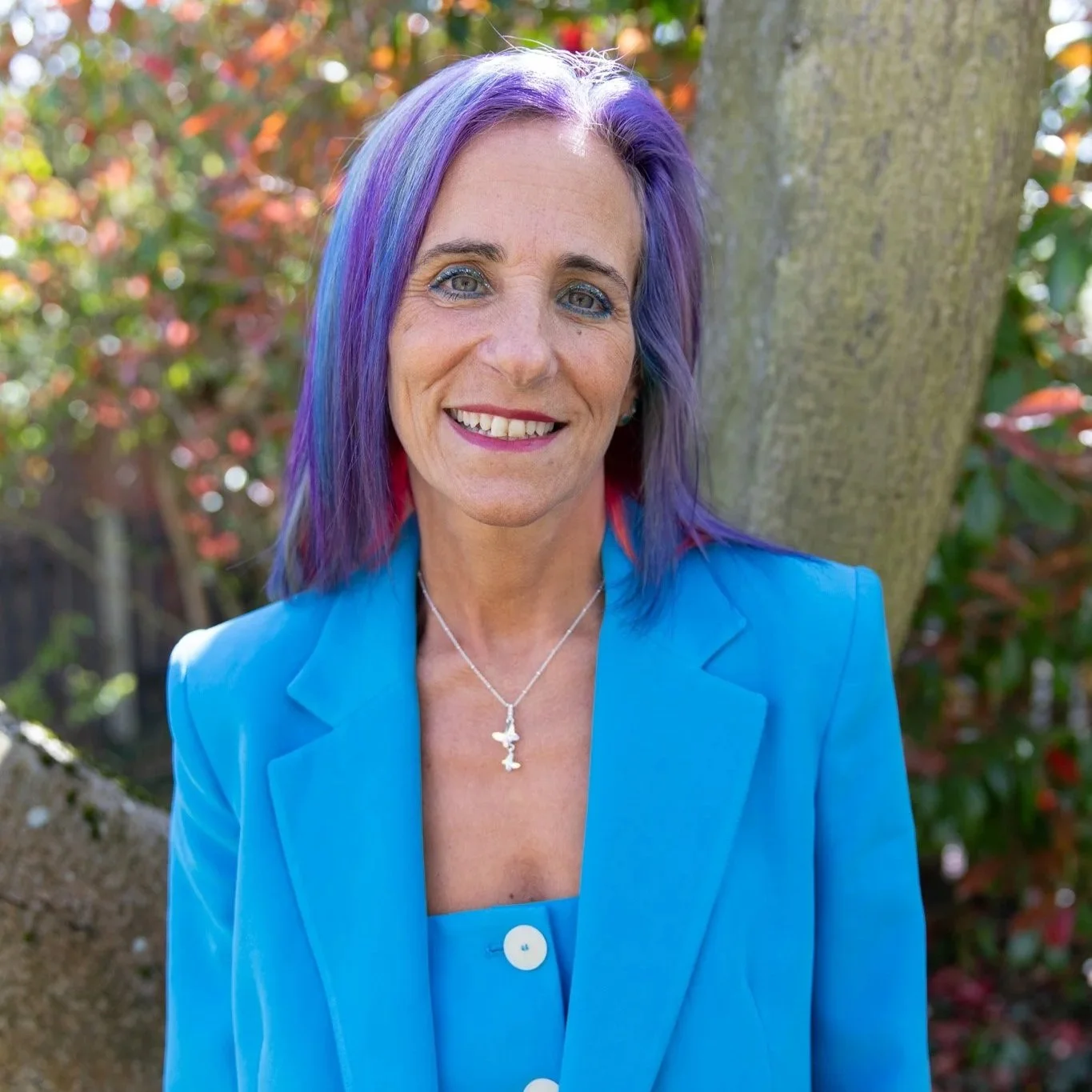 Headshot of Sue Bell, Kids Inspire's CEO and founder. She is outside by a tree and has shoulder length blue and purple hair, wearing a sky blue suit.
