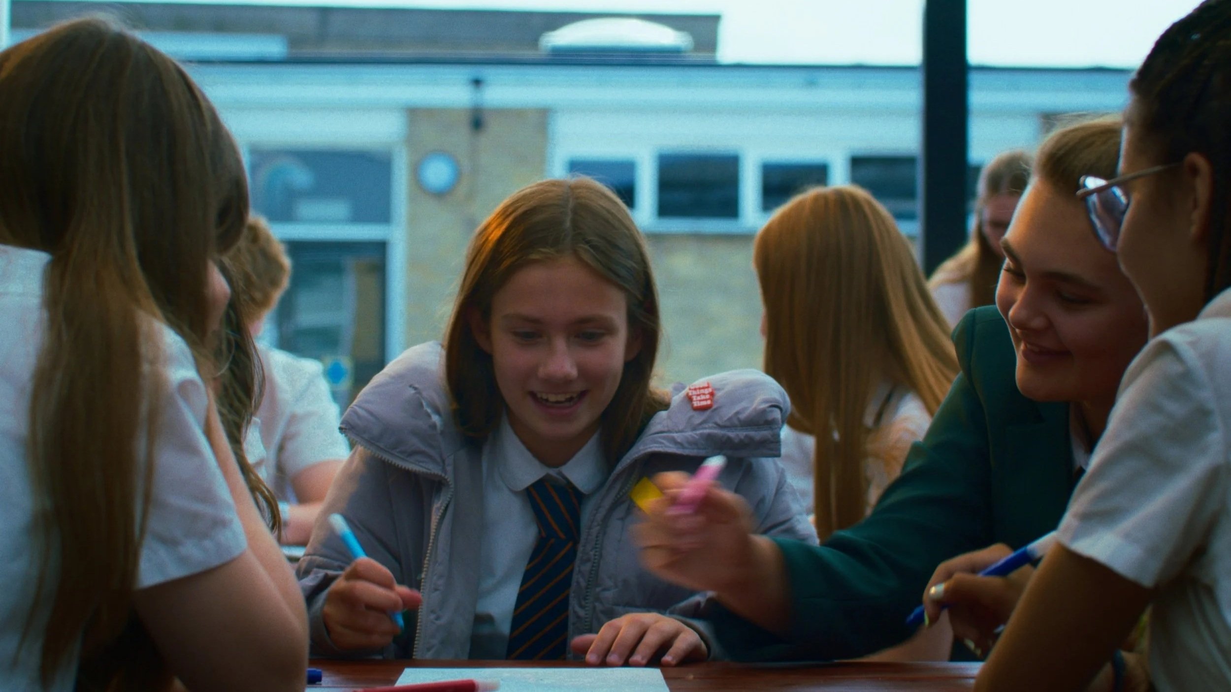 Screengrab of young person sat with friends at lunch, wearing school uniform and oversized coat with coloured tape and badges.