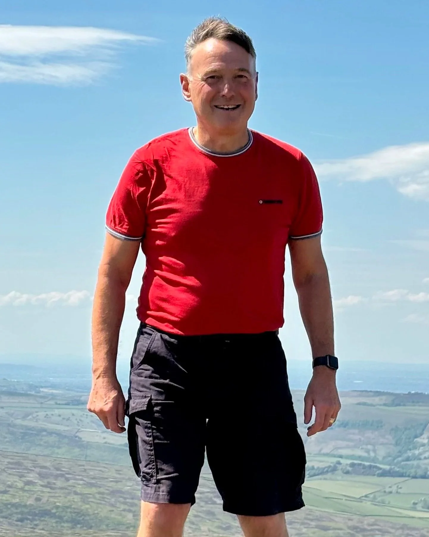 A smiling man with short grey hair, wearing a  red t-shirt and black shorts, stands high up on a mountain with fields of green and blue skies in the background