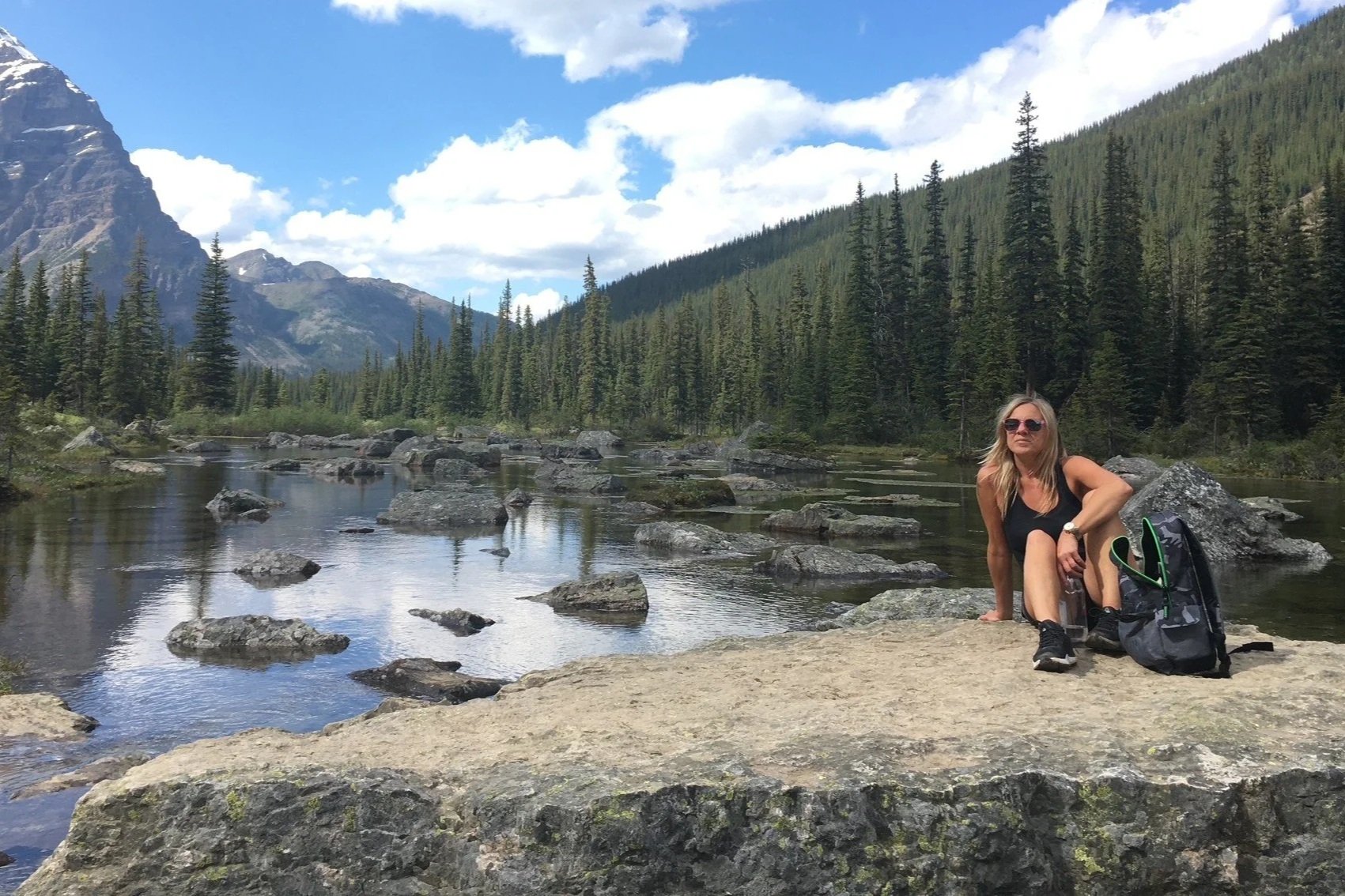 A woman in a vest top and shorts sat by a lake with mountains and blue skies in the background. She is a Kids Inspire volunteer mentor.