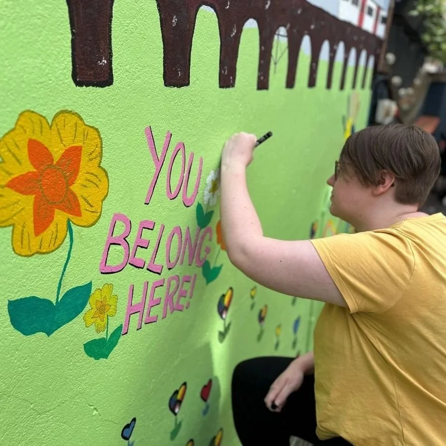 Young person with short hair is drawing on a mural as part of a group project, with the words 'you belong here' included. There are colourful flowers and pride flag hearts too on a green wall.