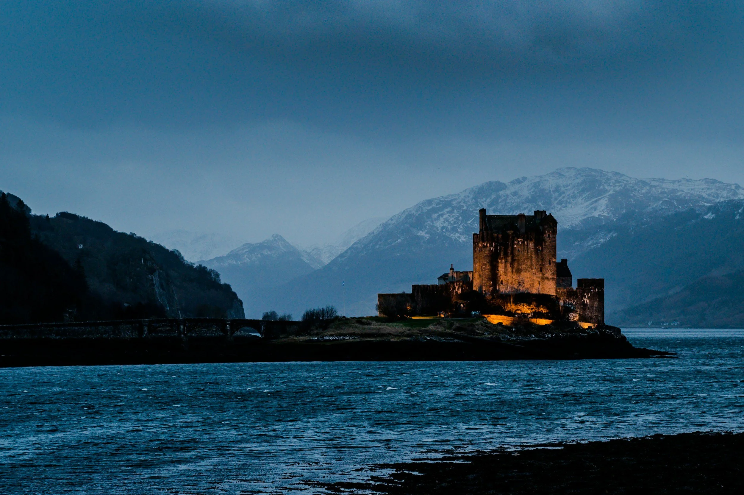 A medieval castle illuminated at dusk on a small island in a body of water, with mountains in the background under a cloudy sky.