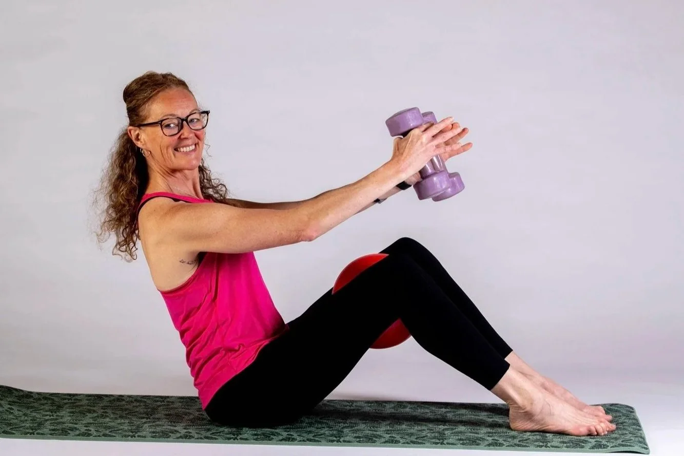 Lynda seated on a mat, knees bent, smiling and holding hand weights
