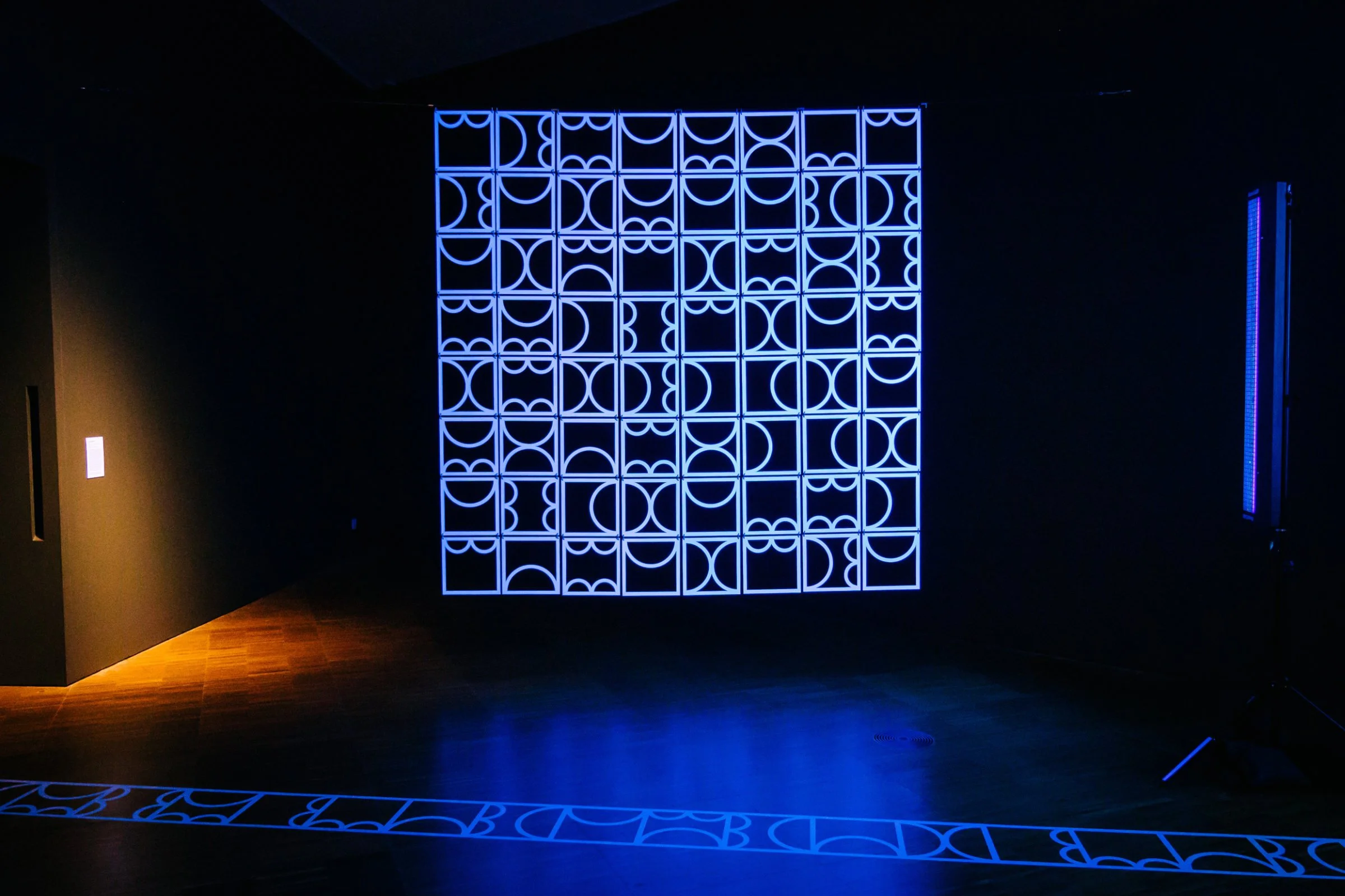 Gallery installation view of shemza.digital #9 glowing under ultraviolet light, with the geometric sculpture reflected across the gallery floor.
