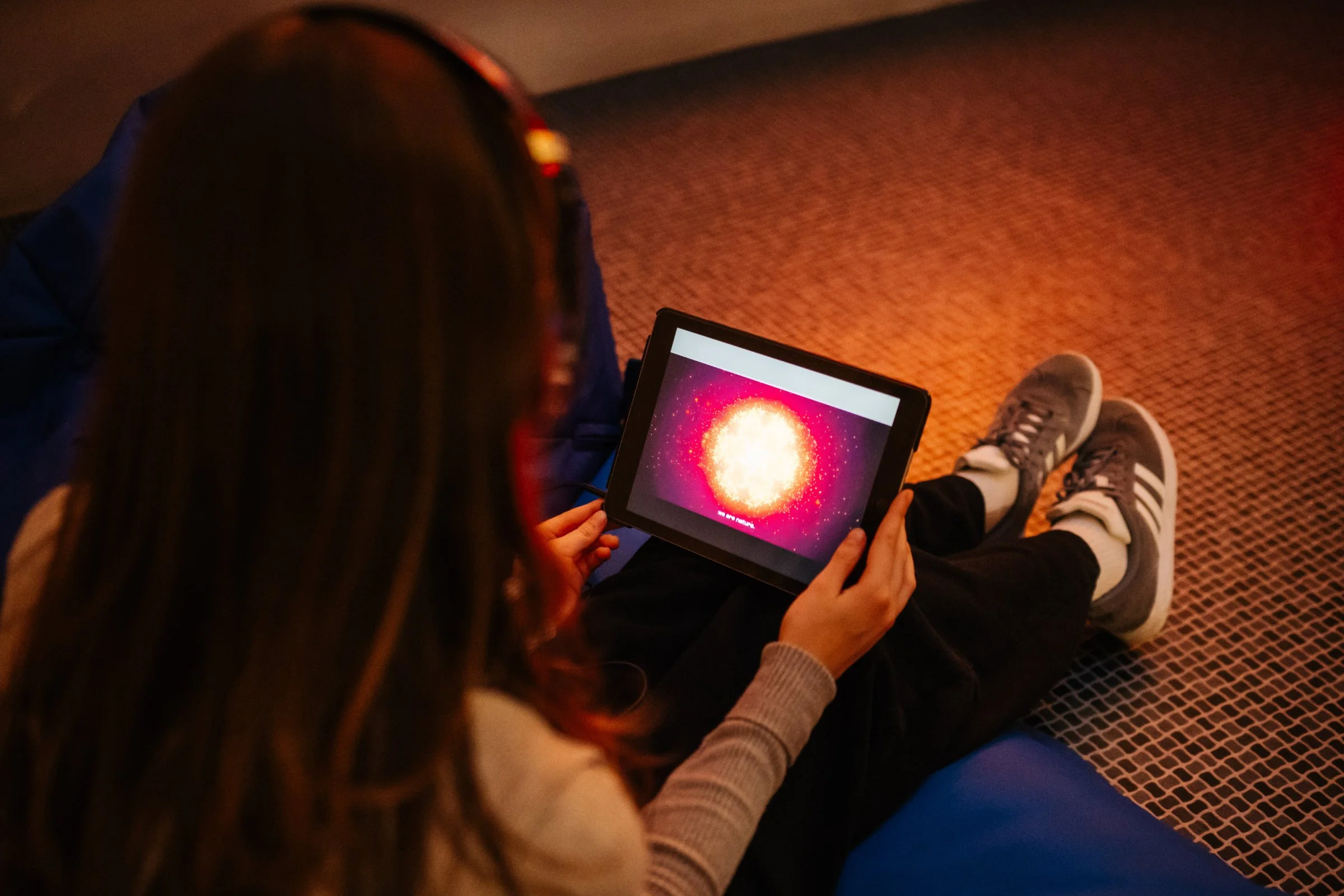 Visitor seated on a beanbag experiencing shemza.digital roots #1 on an iPhone with headphones as glowing red calligraphic forms appear on screen.