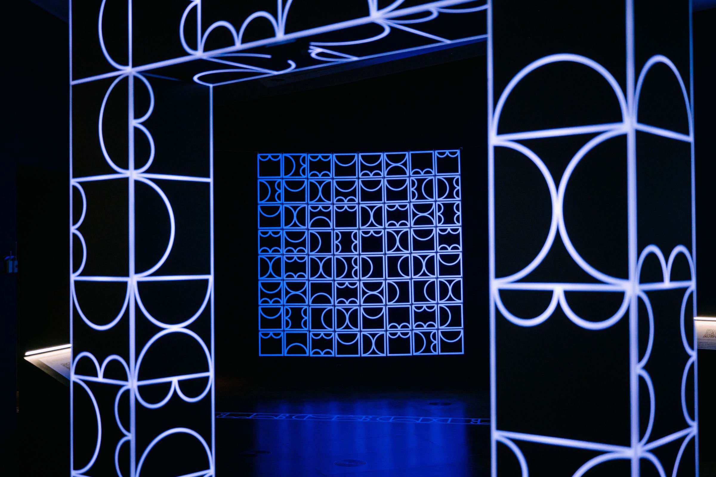 View through an archway of shemza.digital #9 by Aphra Shemza, a suspended ultraviolet light sculpture inspired by Indo-Islamic jali screens and geometric architectural forms.