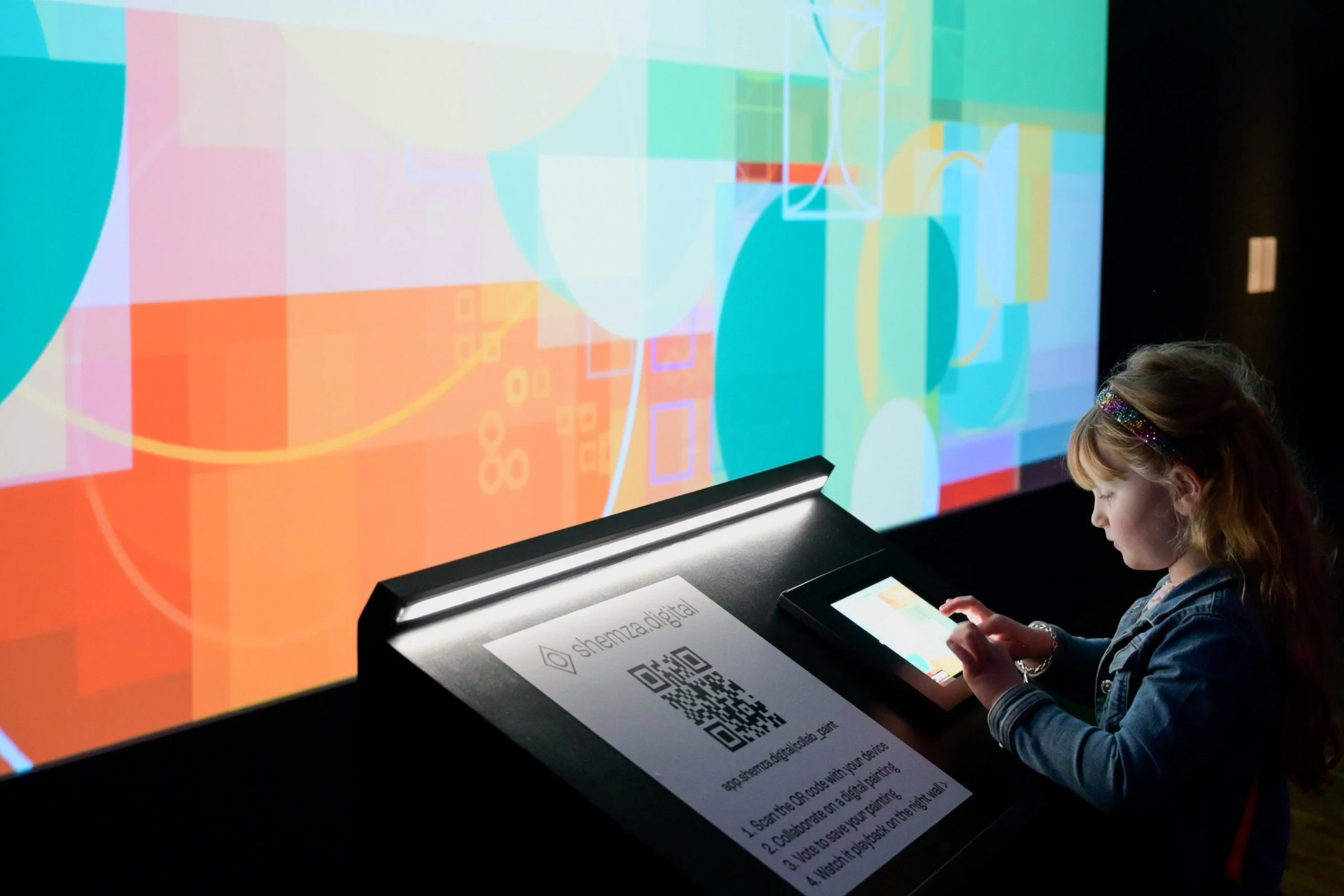 Child creating a collaborative digital painting for shemza.digital #4–7, with the artwork appearing live on the projected gallery wall.