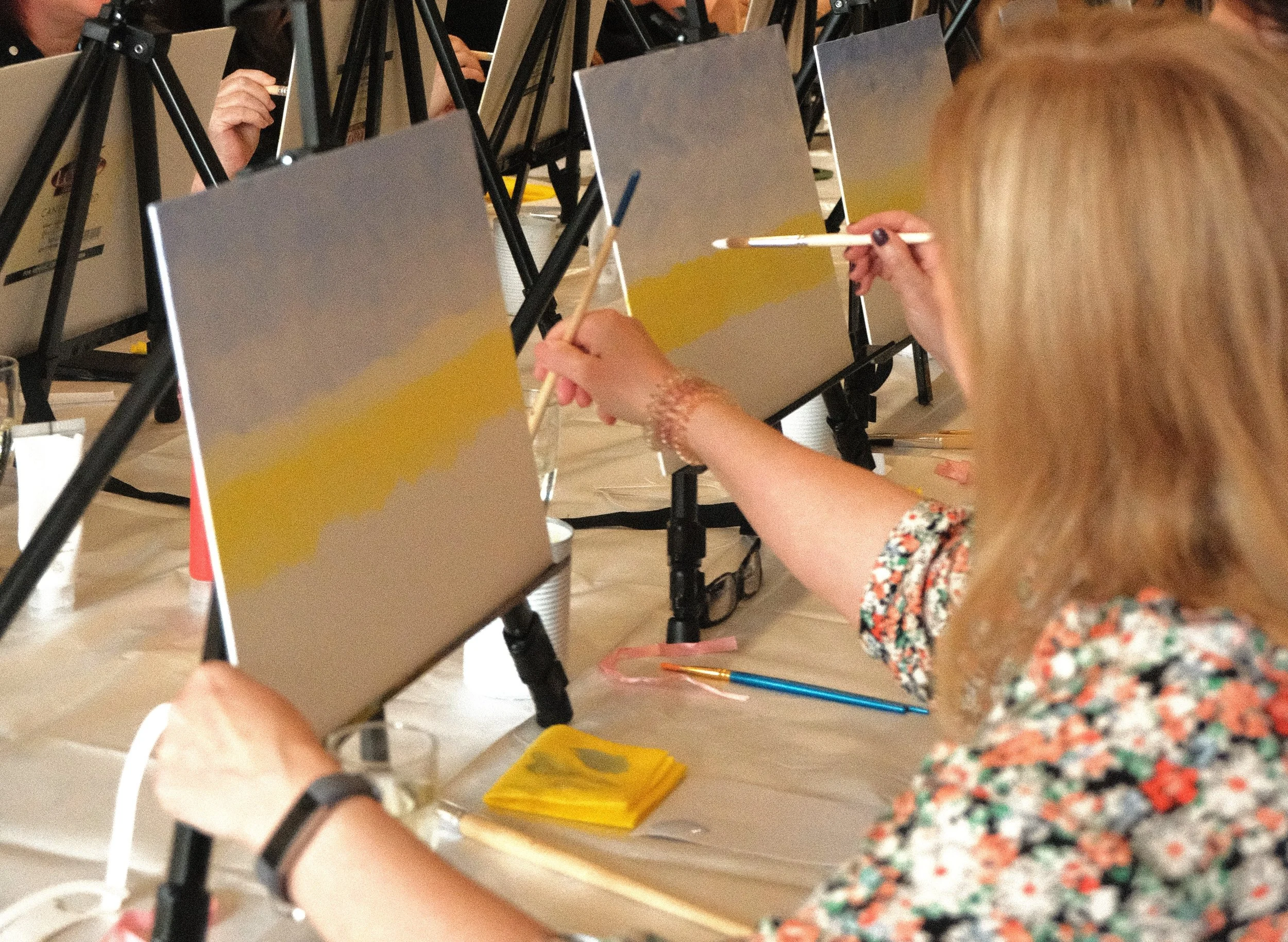 A woman painting a small object at a paint and sip event, with multiple easels and mirrors in the background.
