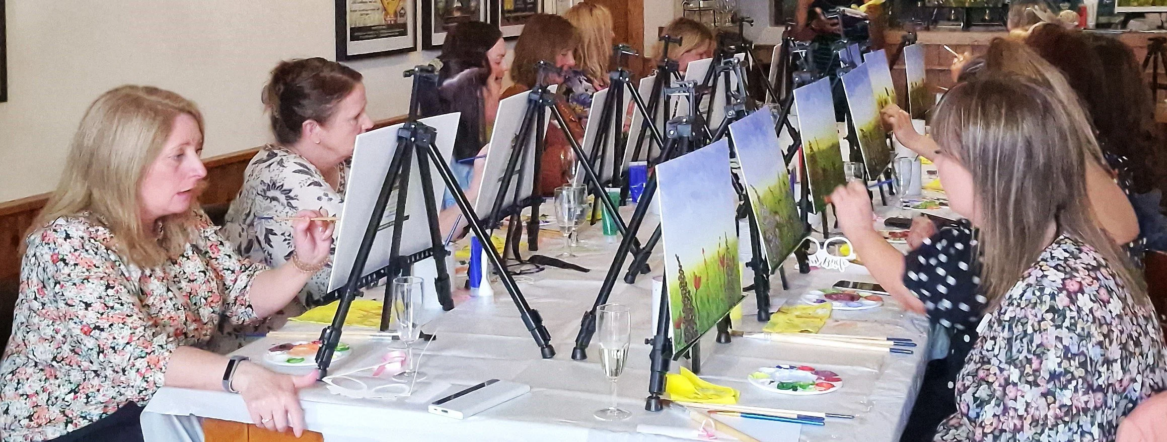 A group of women participating in a painting class, sitting at a long table with easels, paintings, and painting supplies, focusing on their artwork.