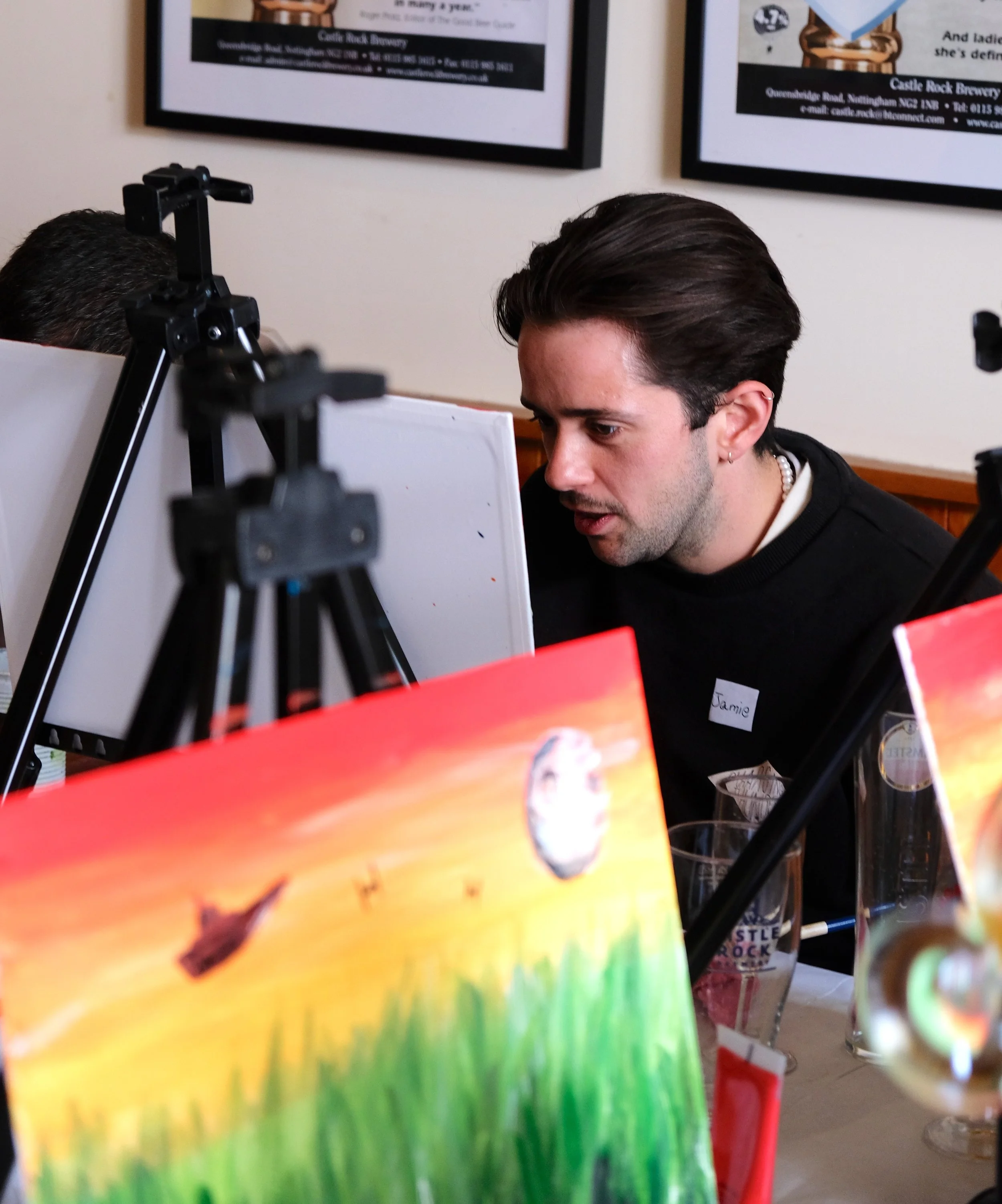Close-up of a colorful rainbow rainbow painting in the foreground with a person with dark hair and a pearl necklace sitting at a table in the background. The person has a name tag that reads 'Jamie'. Two framed posters are hanging on the wall behind 