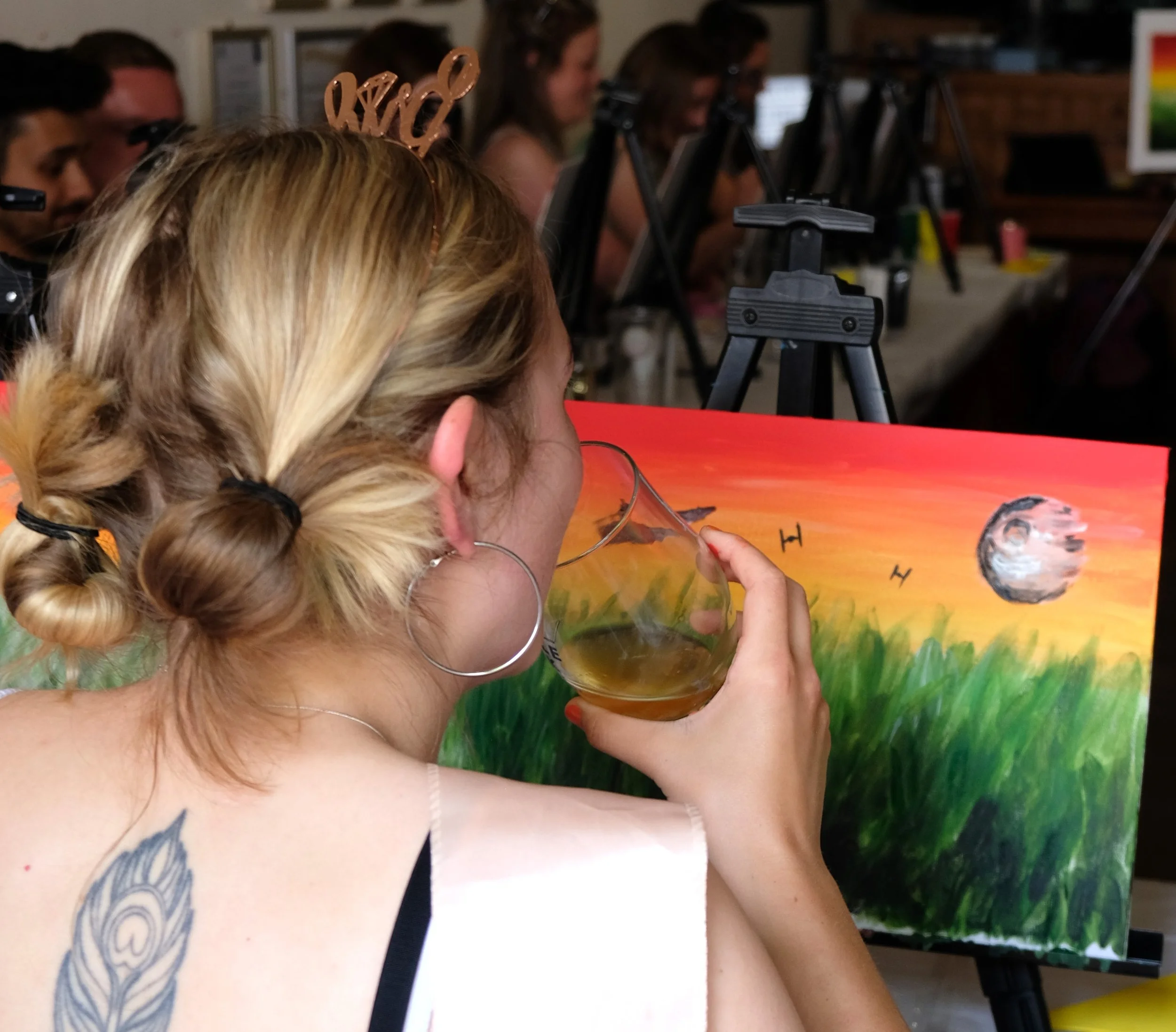 A woman with blonde hair in a braid wearing large hoop earrings, seated at a table, holding a glass of beer, with a colorful rainbow painting of planets in the background.