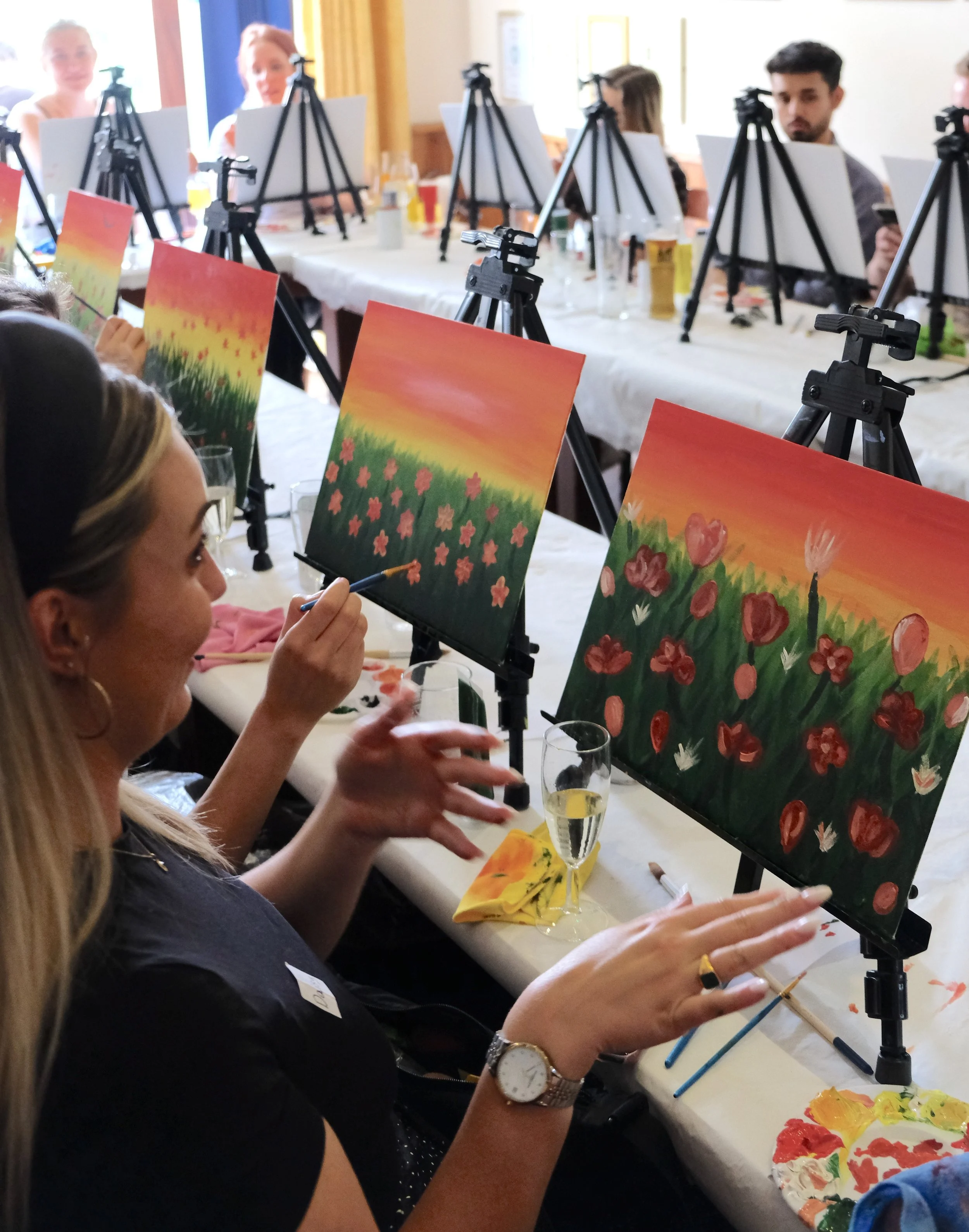 A woman painting at an art class with multiple colorful floral paintings on easels in front of her. A group of people, including a man and women, are seated at a table with painting supplies, with some in the background, and glasses of drinks on the 