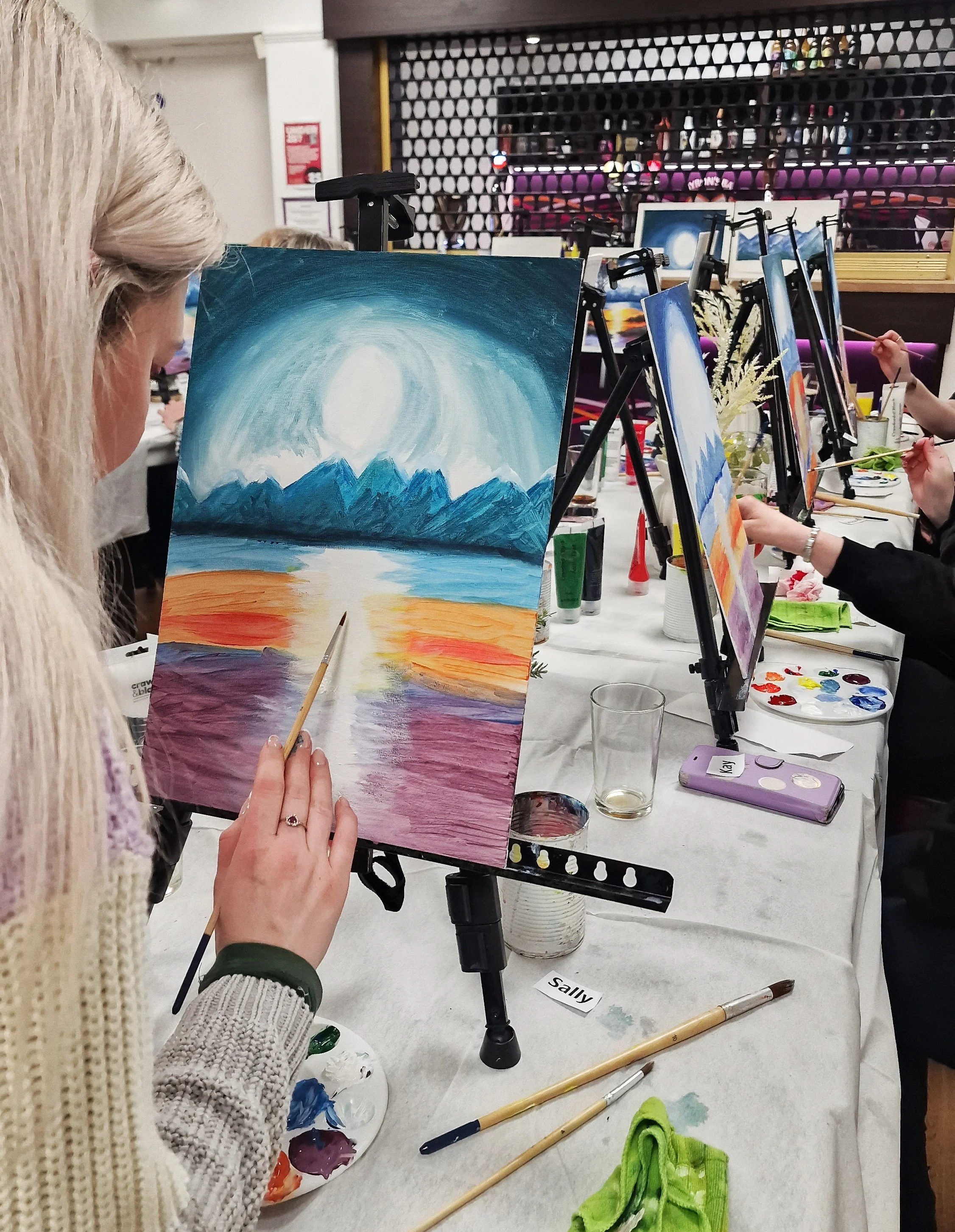 People painting landscapes on easels in an art class, with supplies like paints, brushes, and water on a table.