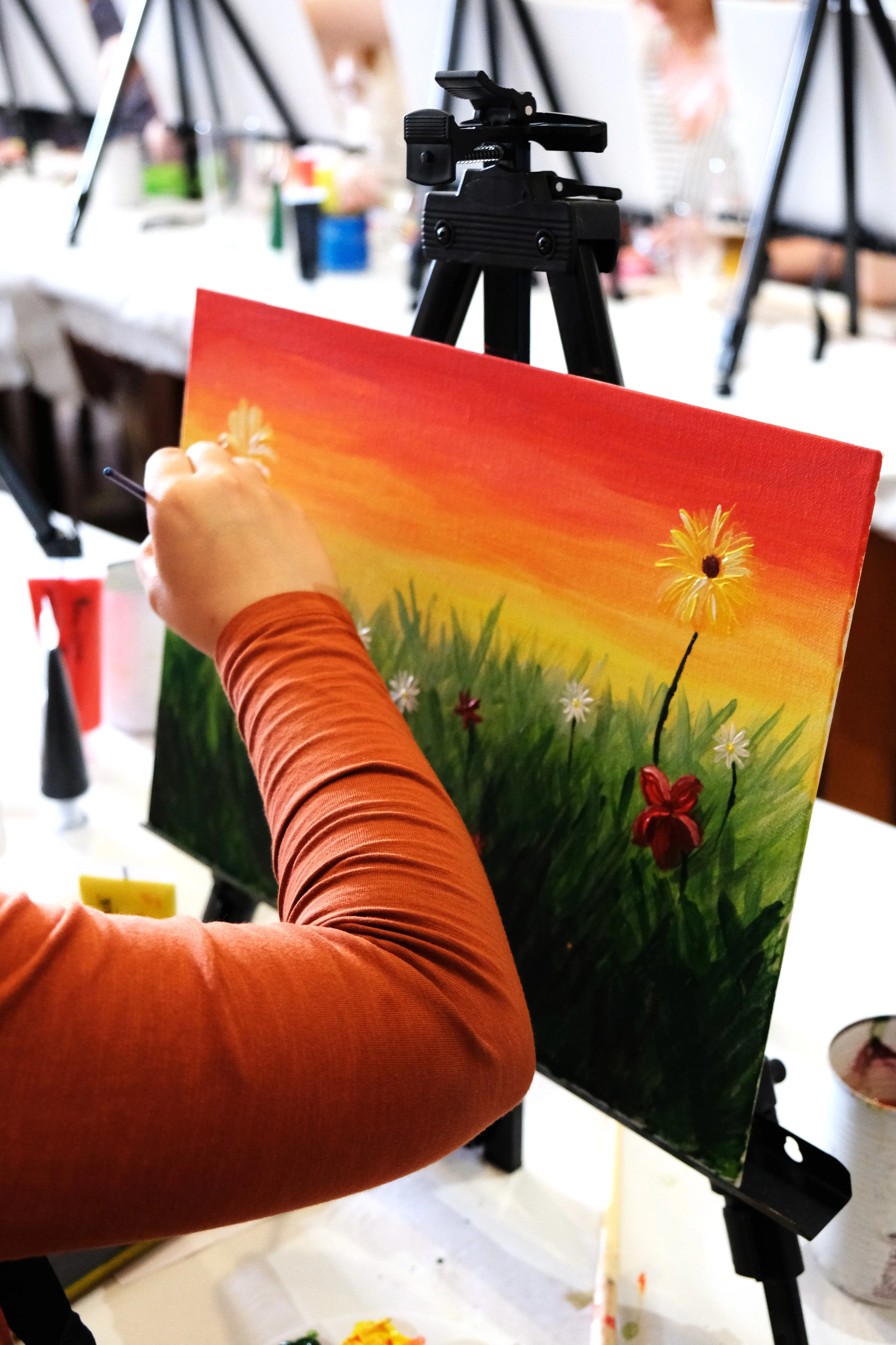 A person wearing an orange long sleeve shirt is painting a colorful landscape on a canvas. The scene includes a vibrant sunset sky with shades of orange, yellow, and red, with white daisies and red flowers in the foreground.