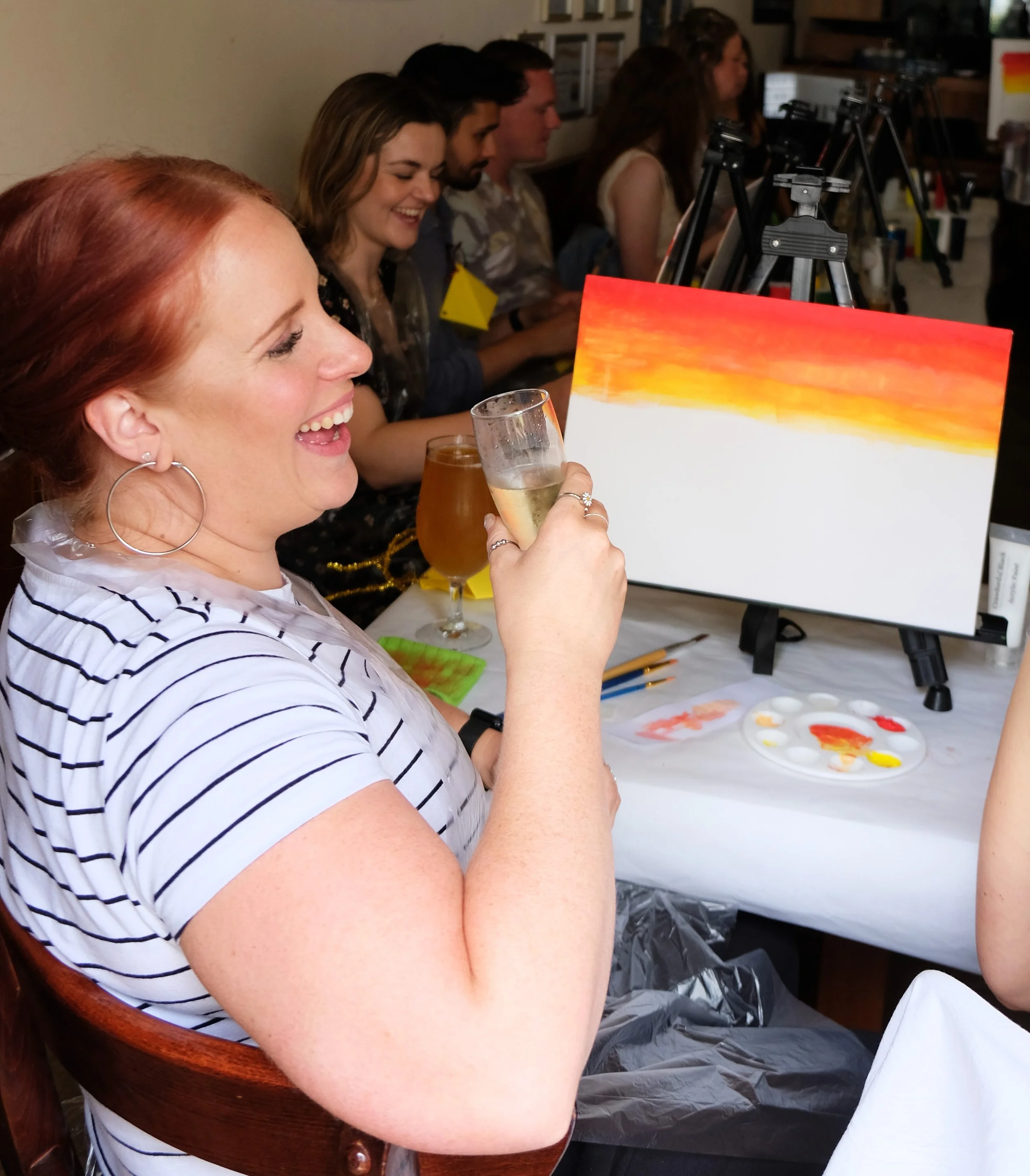 A group of people at an art station, painting and enjoying a social moment. A woman with red hair is smiling and holding a glass of wine, with her eyes closed.