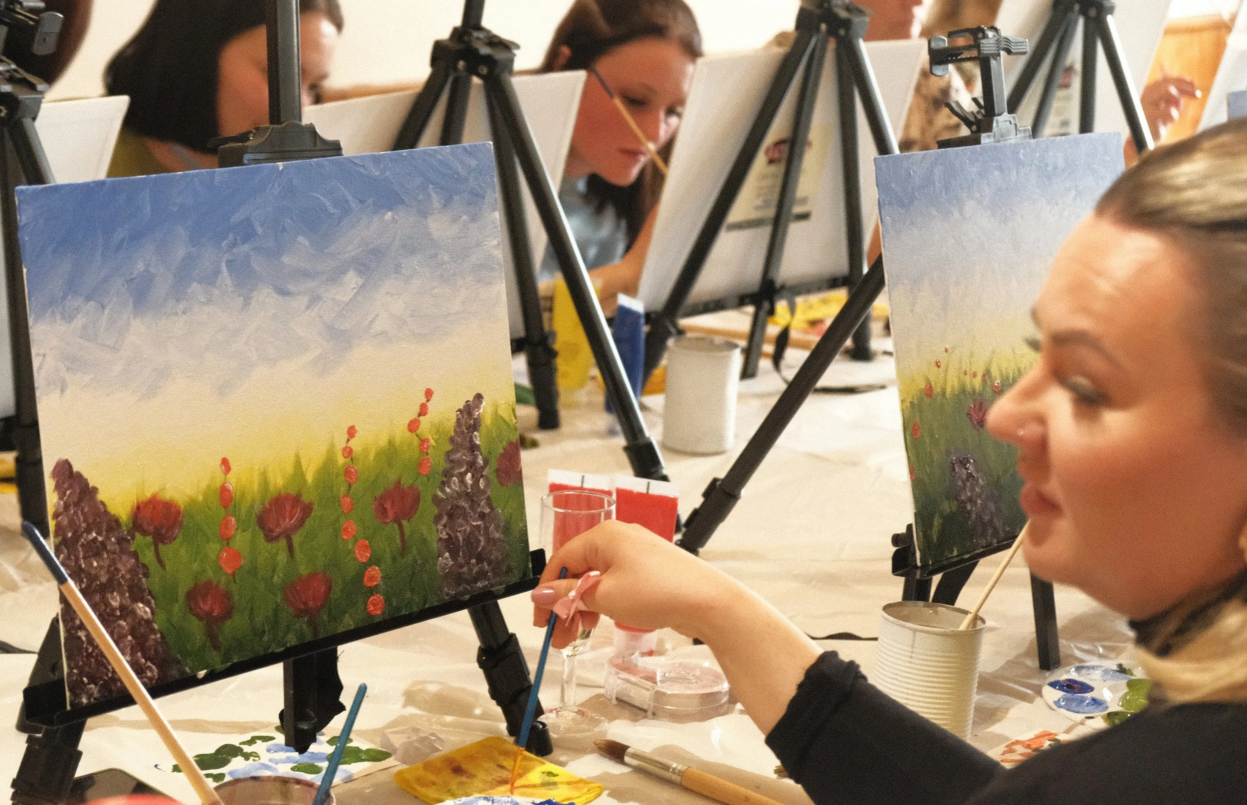 People painting colorful landscape on canvases set up on easels, with focus on a woman painting a field of purple flowers, in an indoor art class.