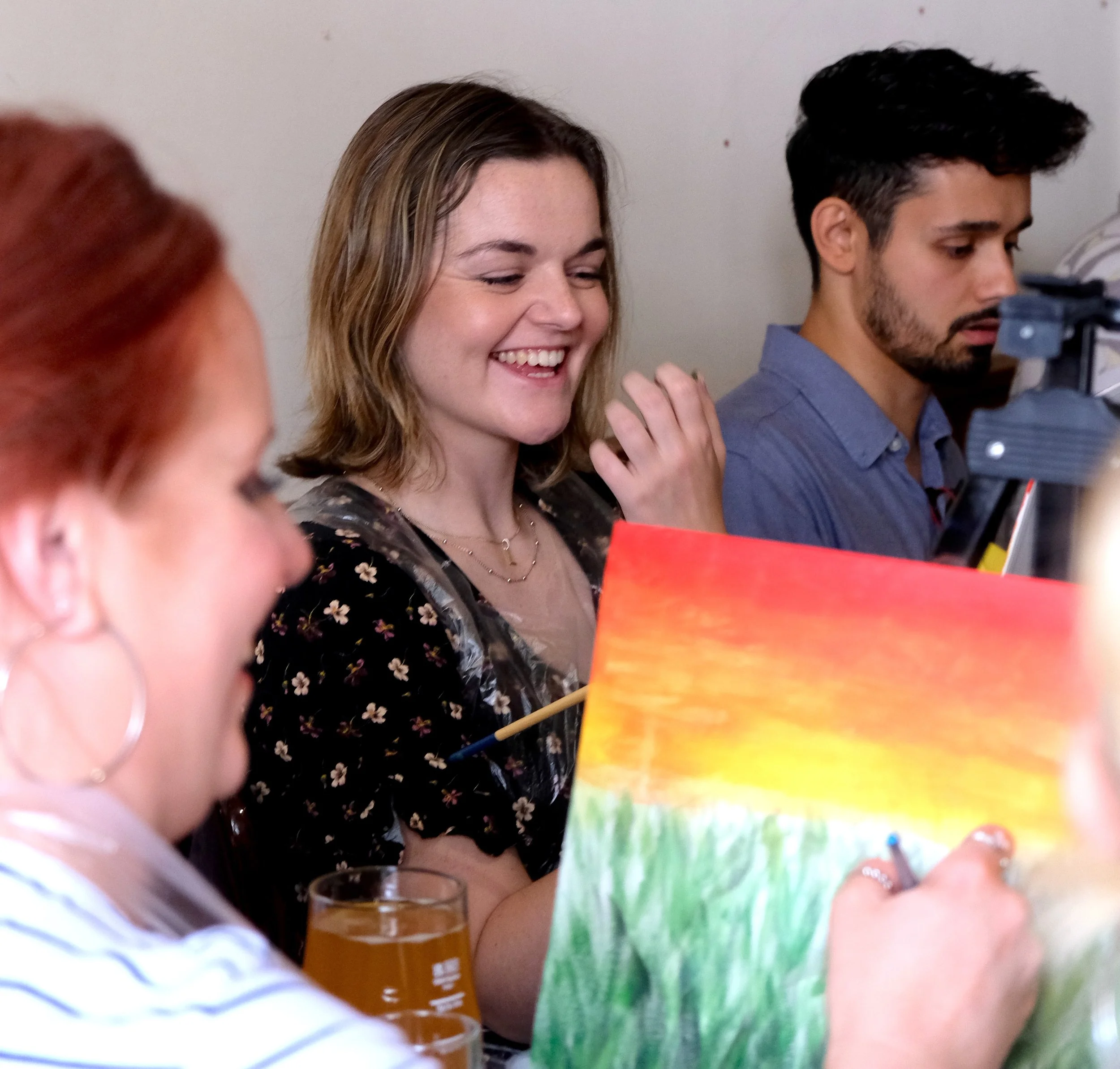 People sitting at a table, with one woman smiling and looking down, while a man on the right is focused on something in front of him. The scene appears to be in a casual setting, possibly a social gathering or a dining environment.
