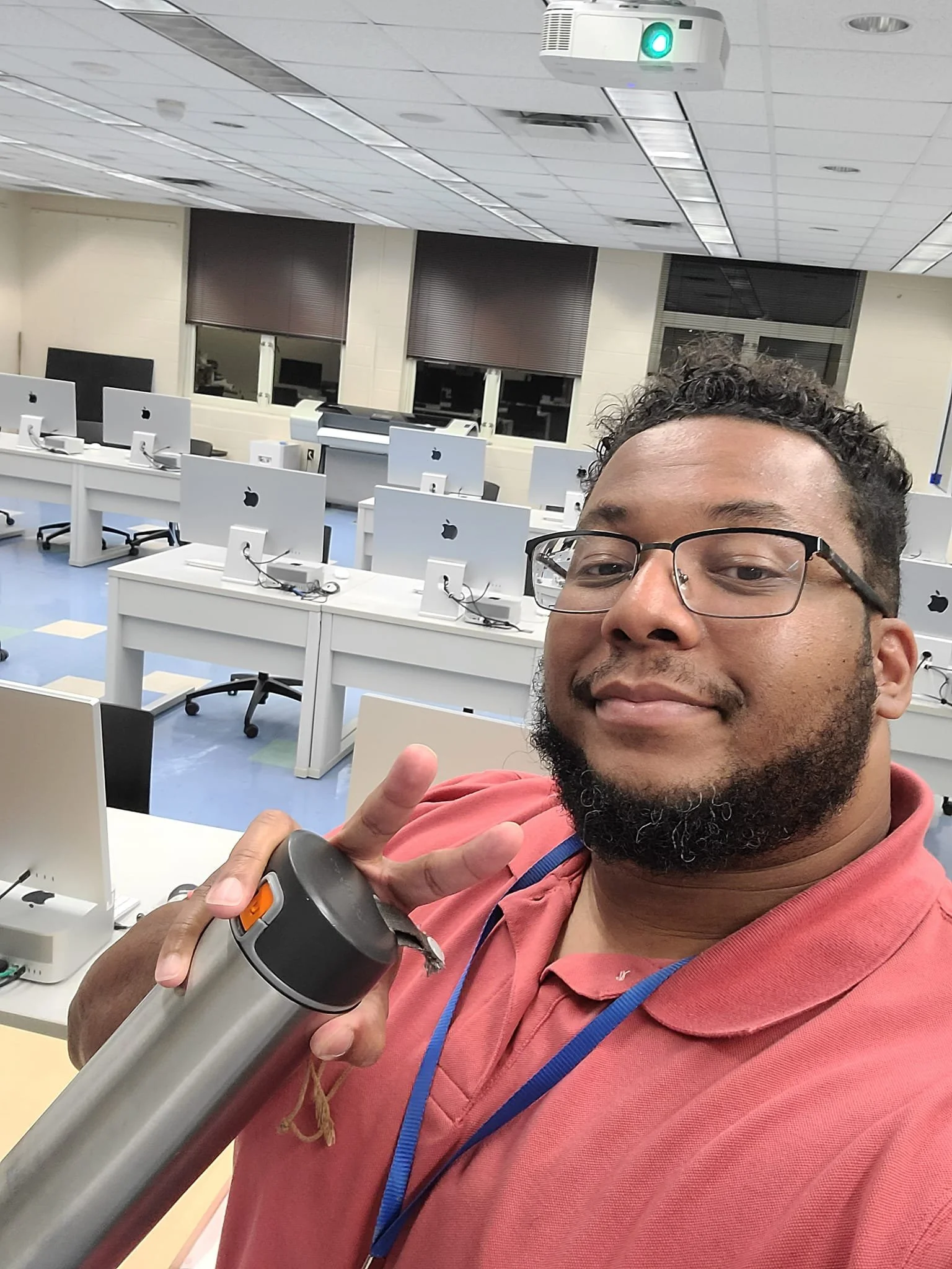 [Image Description: A selfie of a Black man with a beard holding a metal water bottle and smiling. He's wearing glasses and a salmon-colored polo. He's standing in front of a classroom full of Mac computers.]