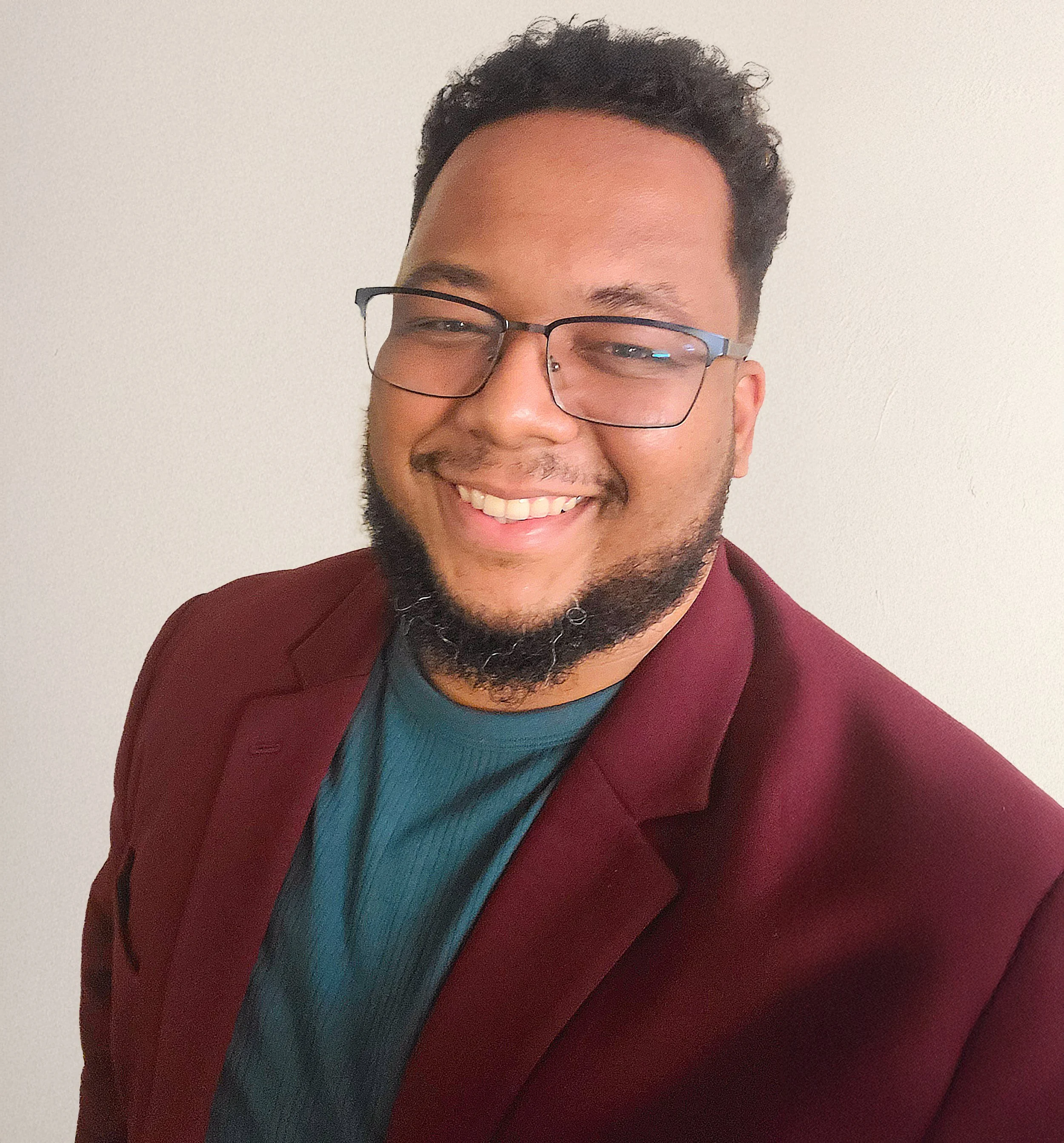 [Image Description: A Black man with a beard and curly hair facing forward and smiling. He's wearing a forest green shirt with a burgundy blazer in front of a neutral background.]