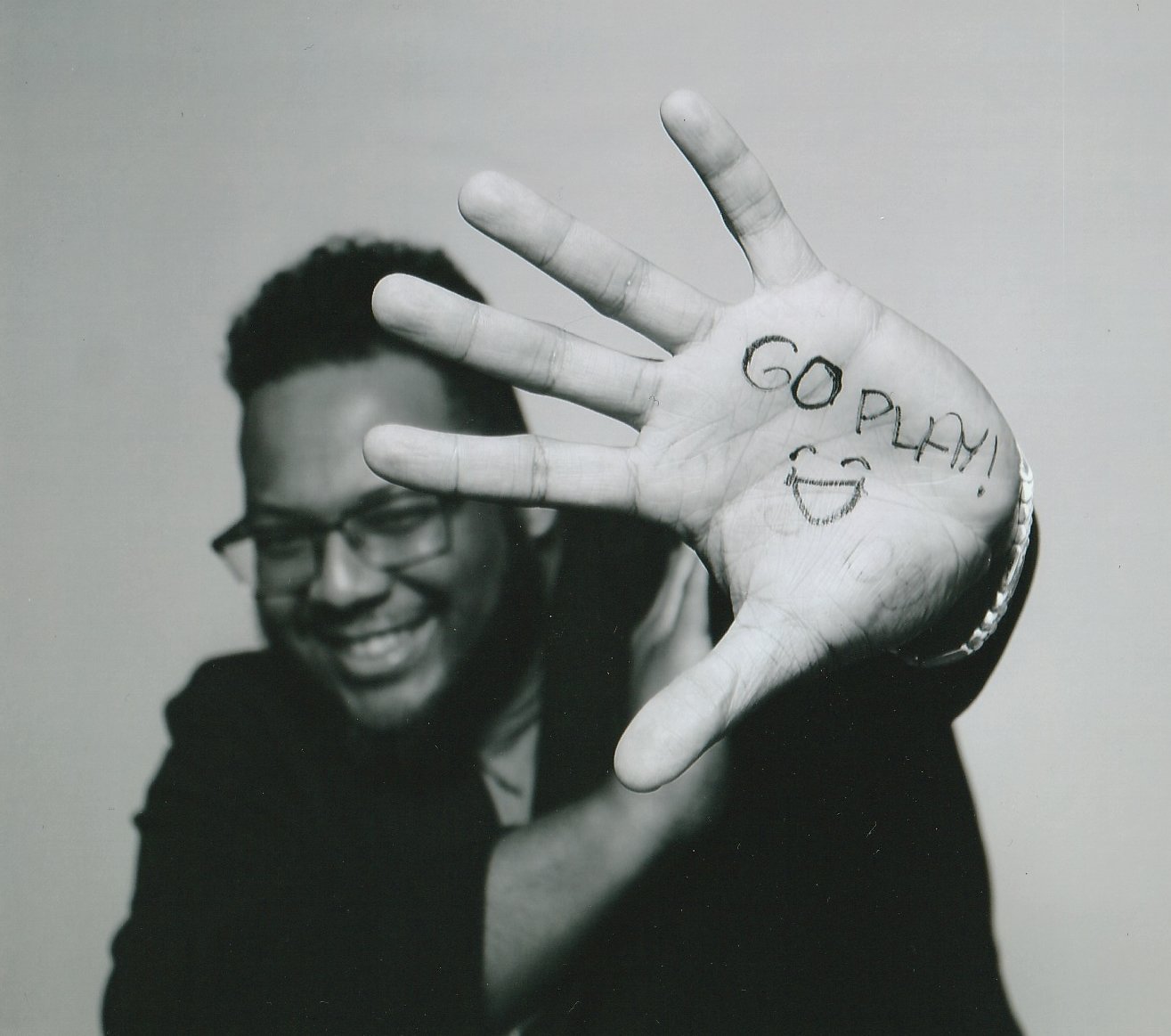 [Image Description: Close-up image of a Black man smiling and holding his hand open towards the camera. "Go play!" with a smiley face is drawn on his palm.]