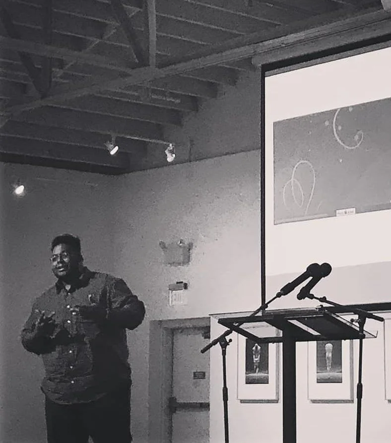 [Image Description: Black and white image of a Black man speaking on stage with graphics on a projector screen behind him.]