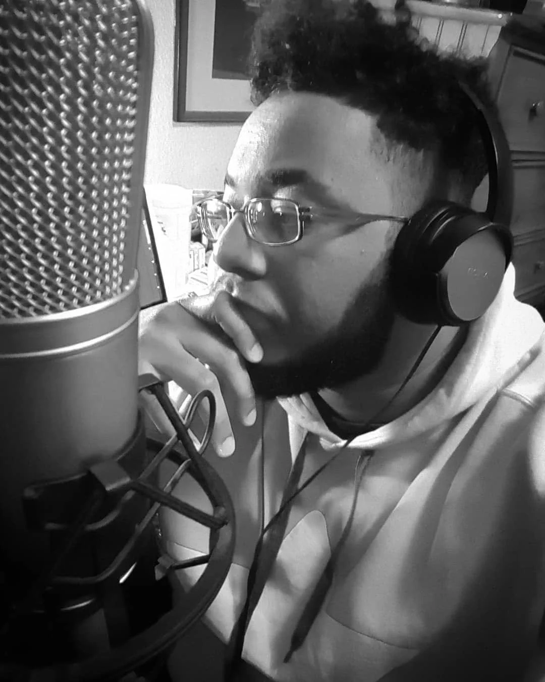 [Image Description: Black and white close-up of a Black man with a high-top fade, beard, glasses, and studio headphones. He's facing left in a contemplative pose with a studio microphone in front of him.]