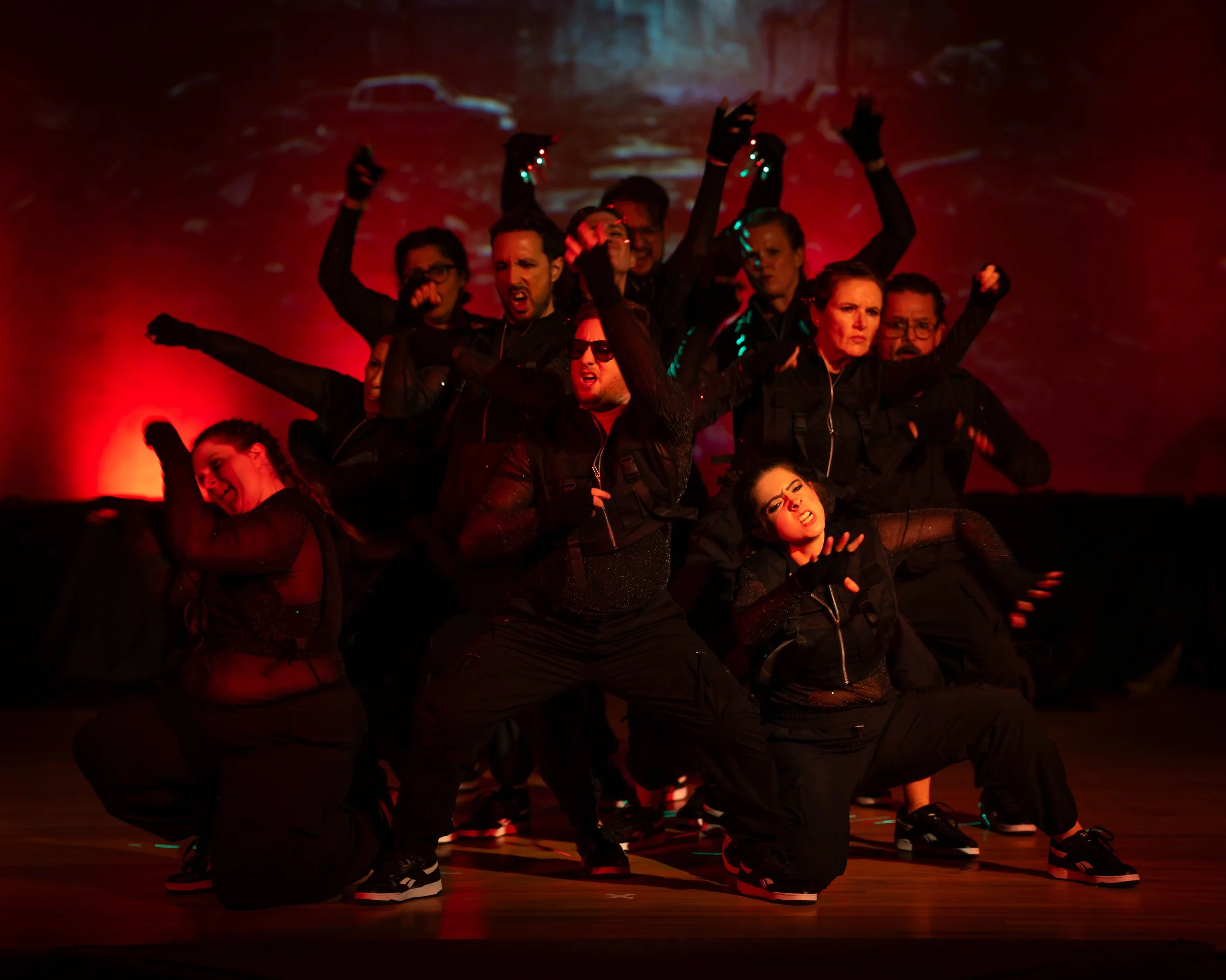 A group of ten dancers performing on stage under red lighting, dressed in black outfits, striking dynamic poses with serious and intense facial expressions.