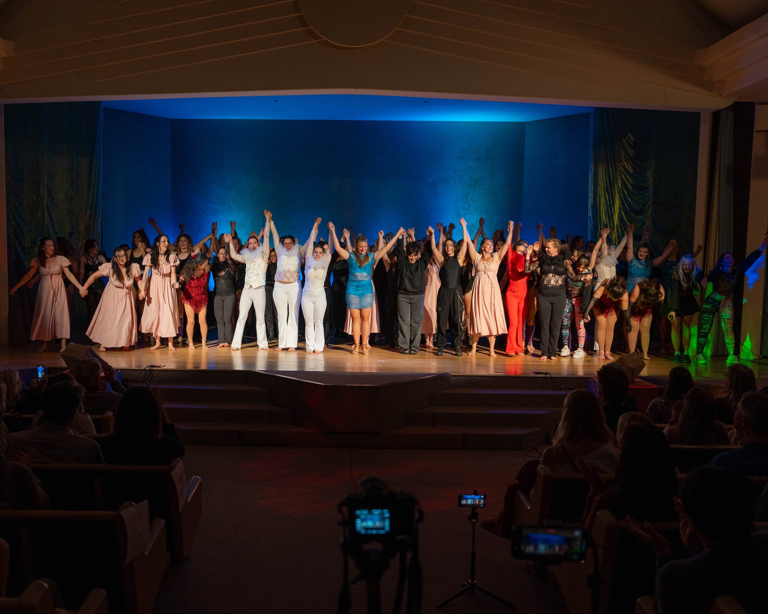 A large group of performers on stage after a show, holding hands and bowing to the audience in a theater with colorful lighting.